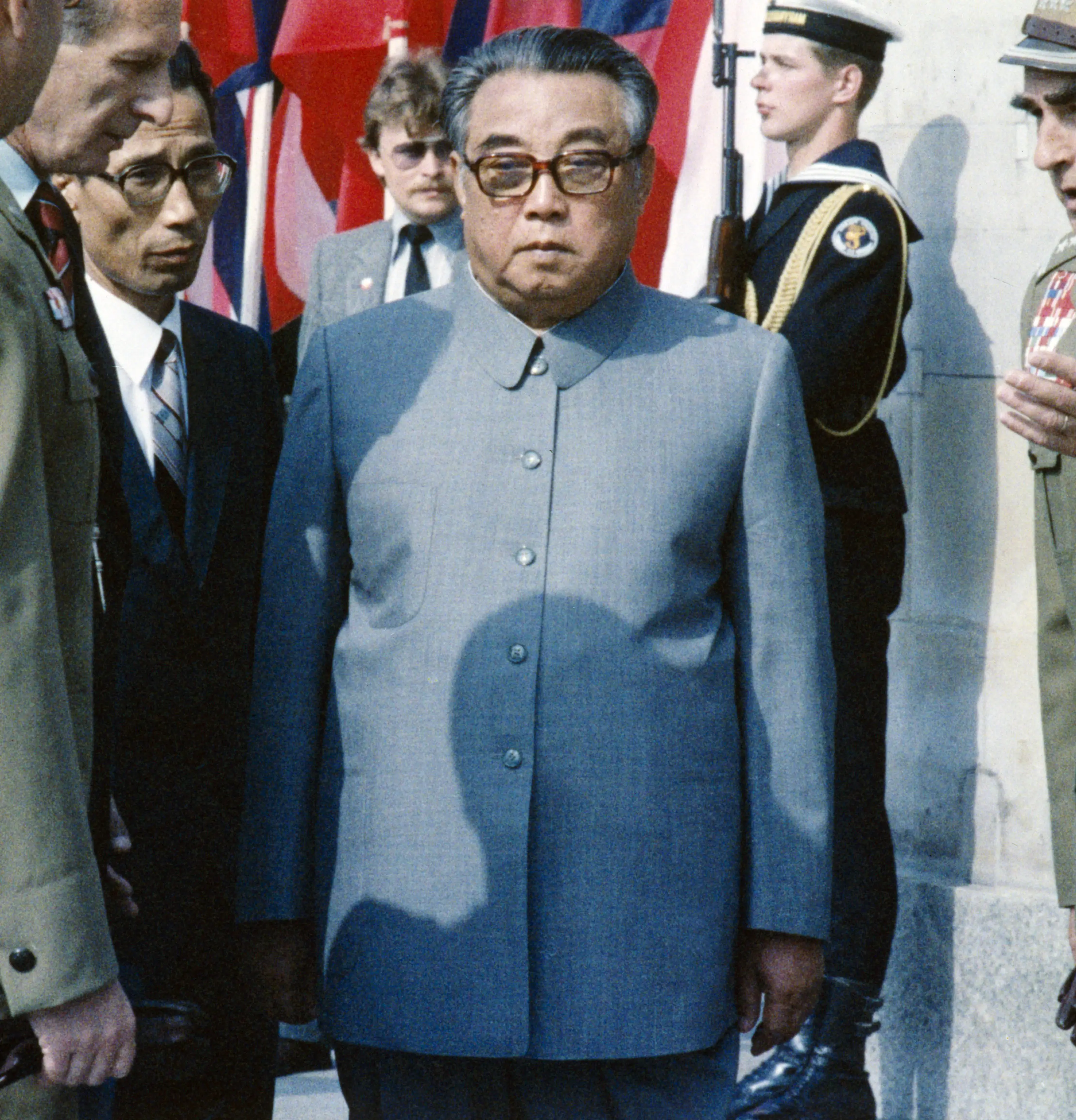 Kim Il Sung wears a gray suit and glasses surrounded by men in suits and military dress