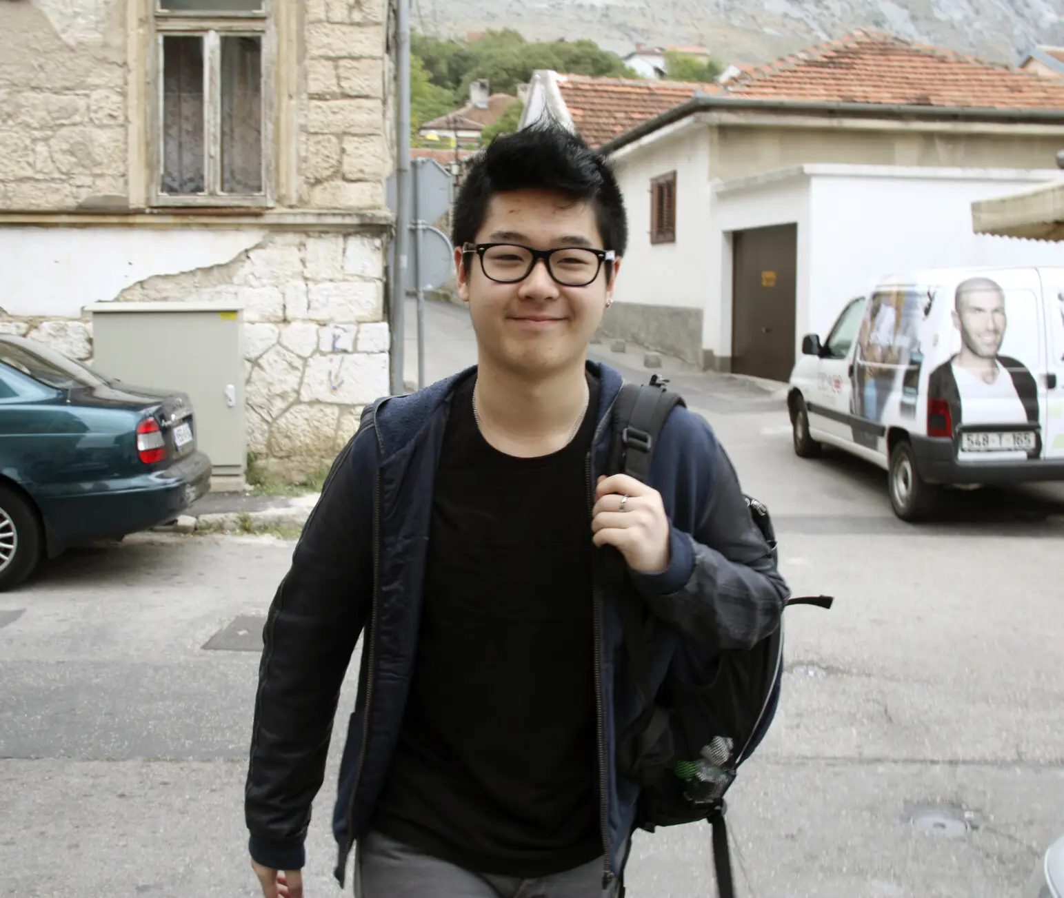 Kim Han Sol smiles with a backpack over one shoulder and small houses and cars behind him on a street