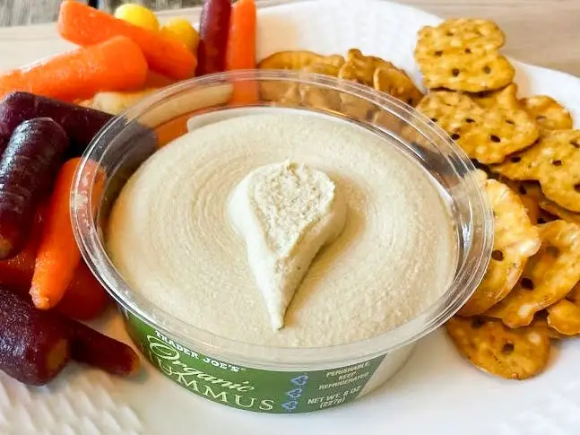 A container of Trader Joe's organic hummus on a plate with multicolored carrots and pretzel crisps.