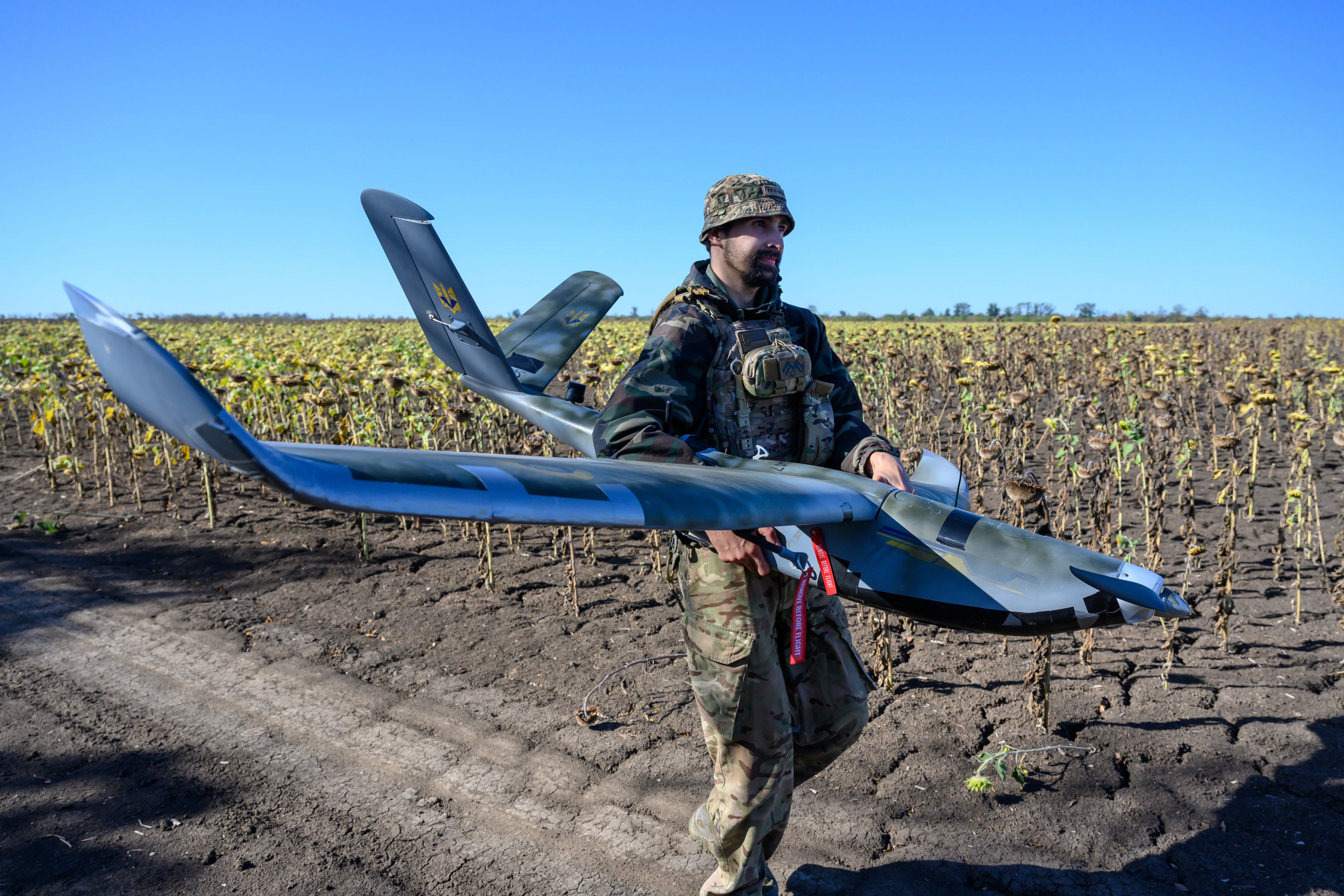 A man walks along a dirt path holding a large drone with a blue sky and crops behind him