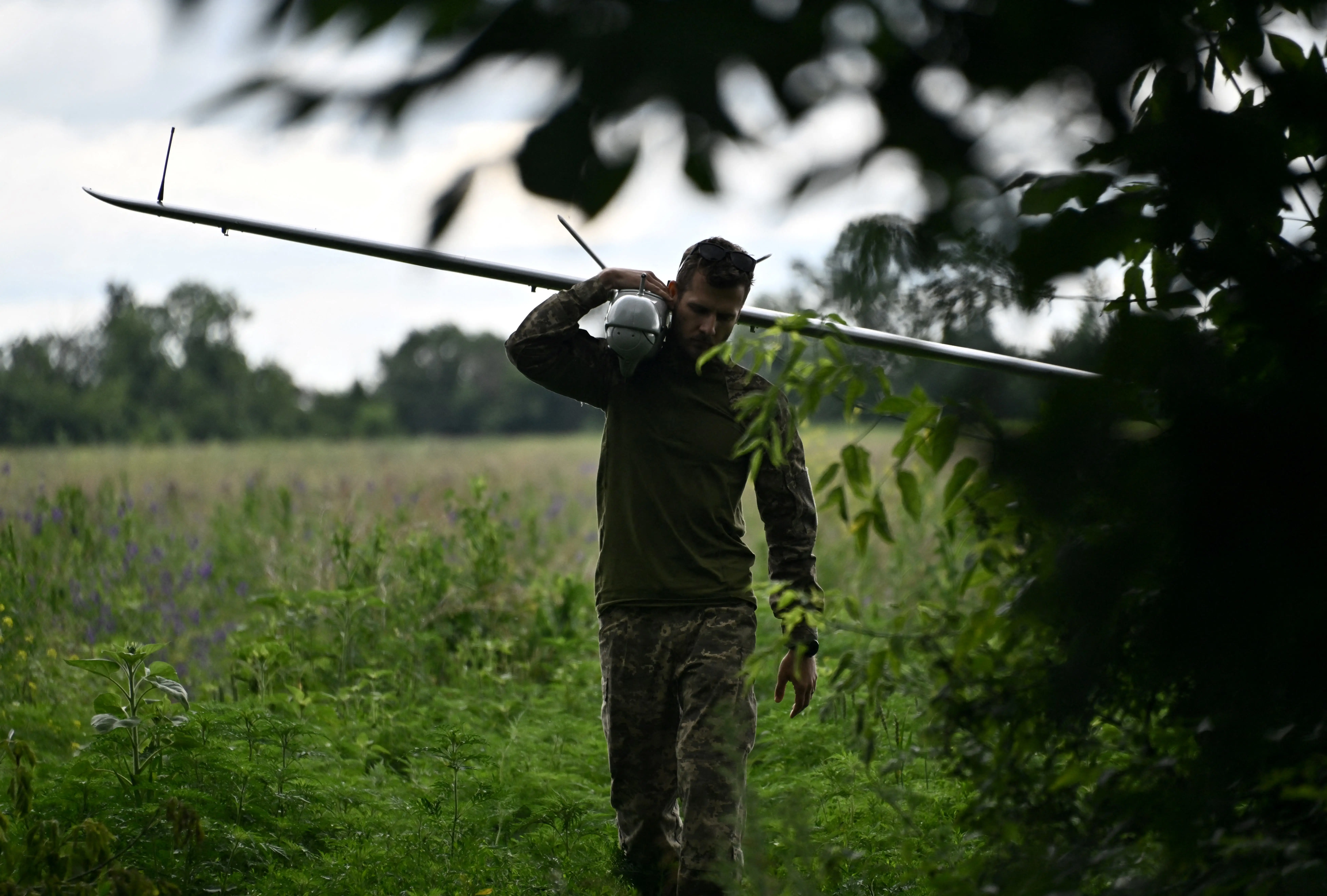 A man in green gear holds a large grey drone over his shoulder as he walks through a green field with leaves in the foreground.