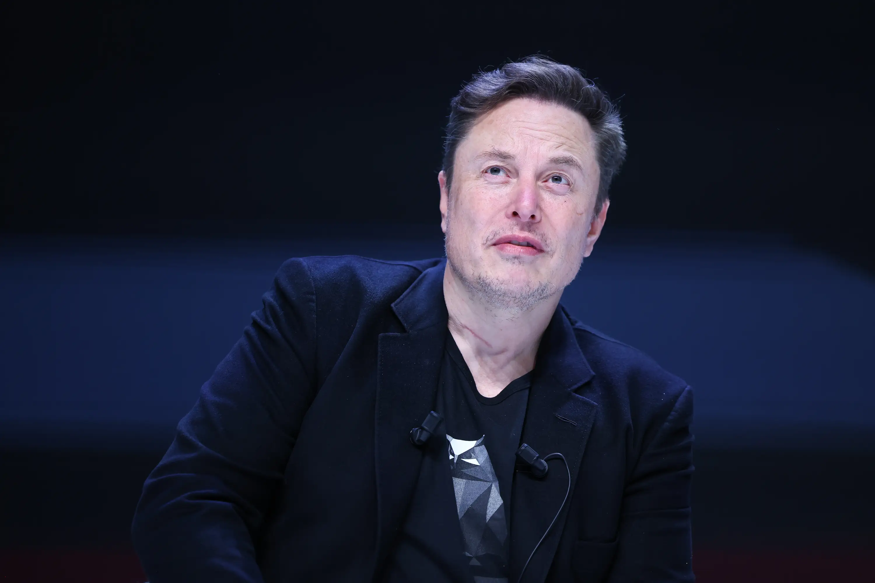 Elon Musk is in a black suit jacket and a black graphic t-shirt on stage. He is looking to the top left corner of the image.