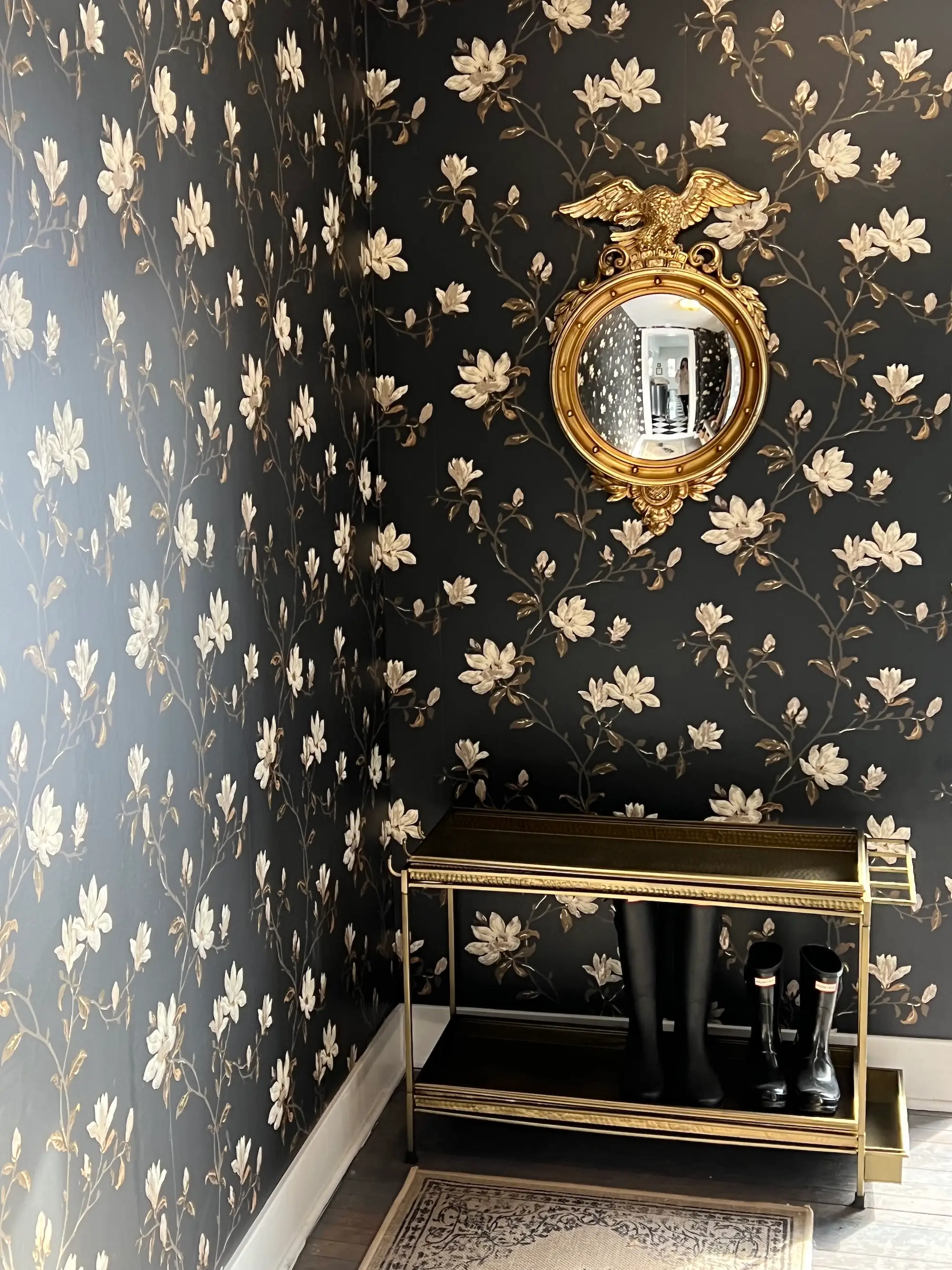 A mudroom with black and white floral walls, a gold mirror, and a shelf for shoes.