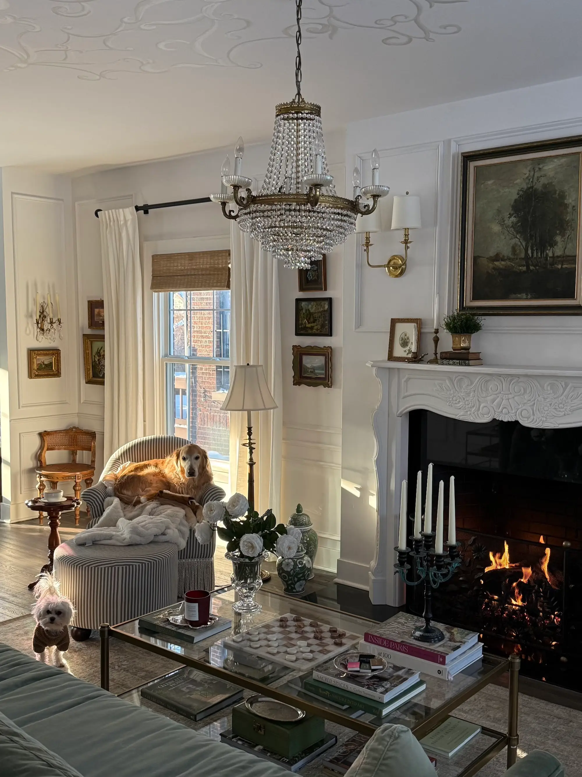 A living room with a white fireplace, white walls, paintings on the walls, and statement chandeliers.