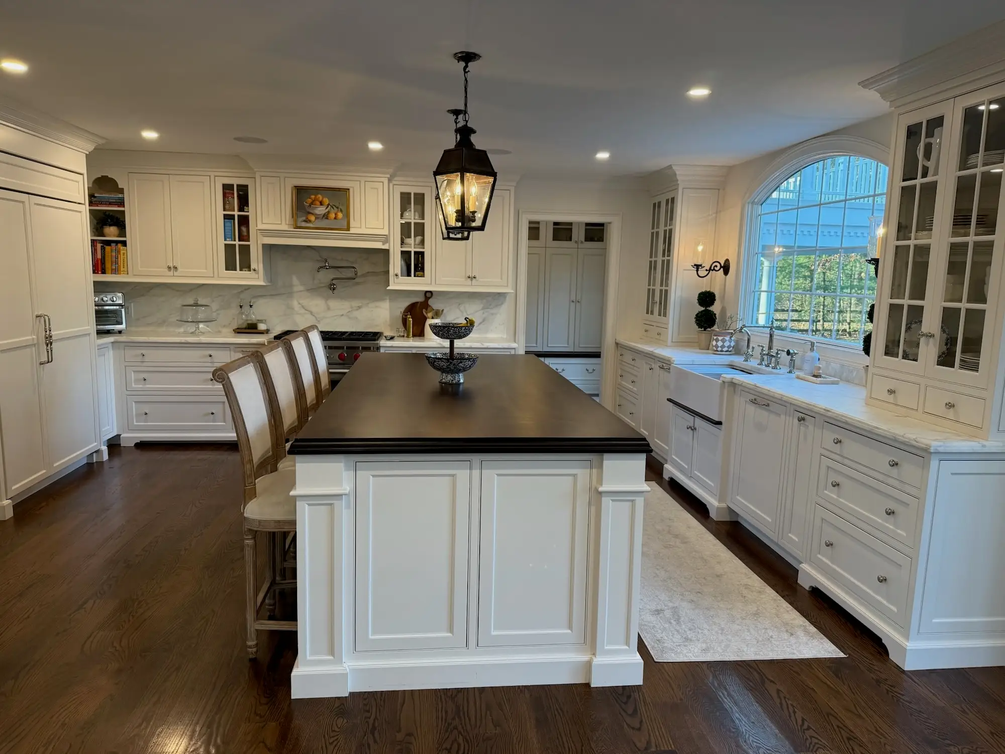 A kitchen with a large wooden island and white cabinetry. An arced window sits atop the sink.