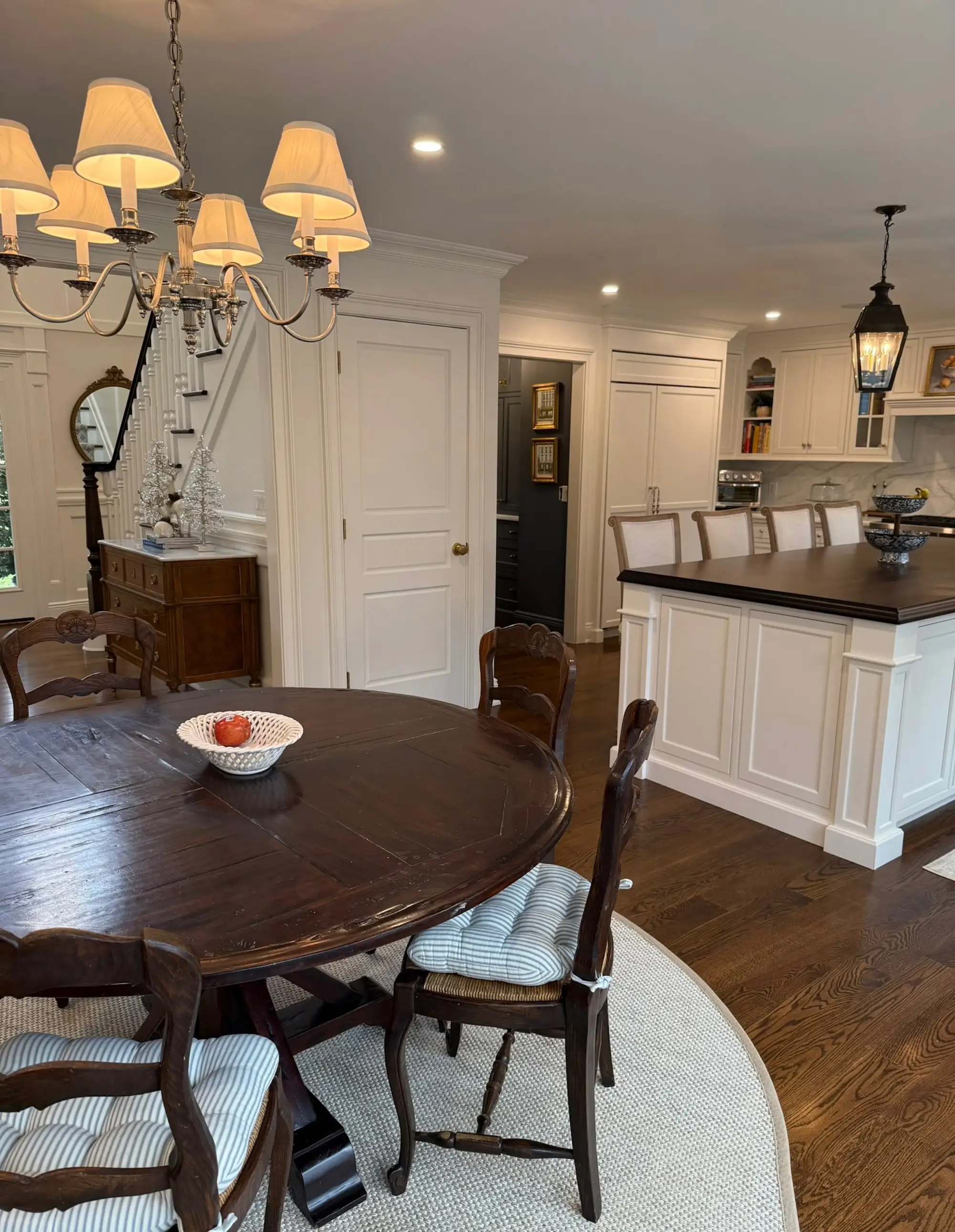 A kitchen with a round, dark dining table and a large chandelier above it. An island sits ot the right with white cabinetry lining the walls.