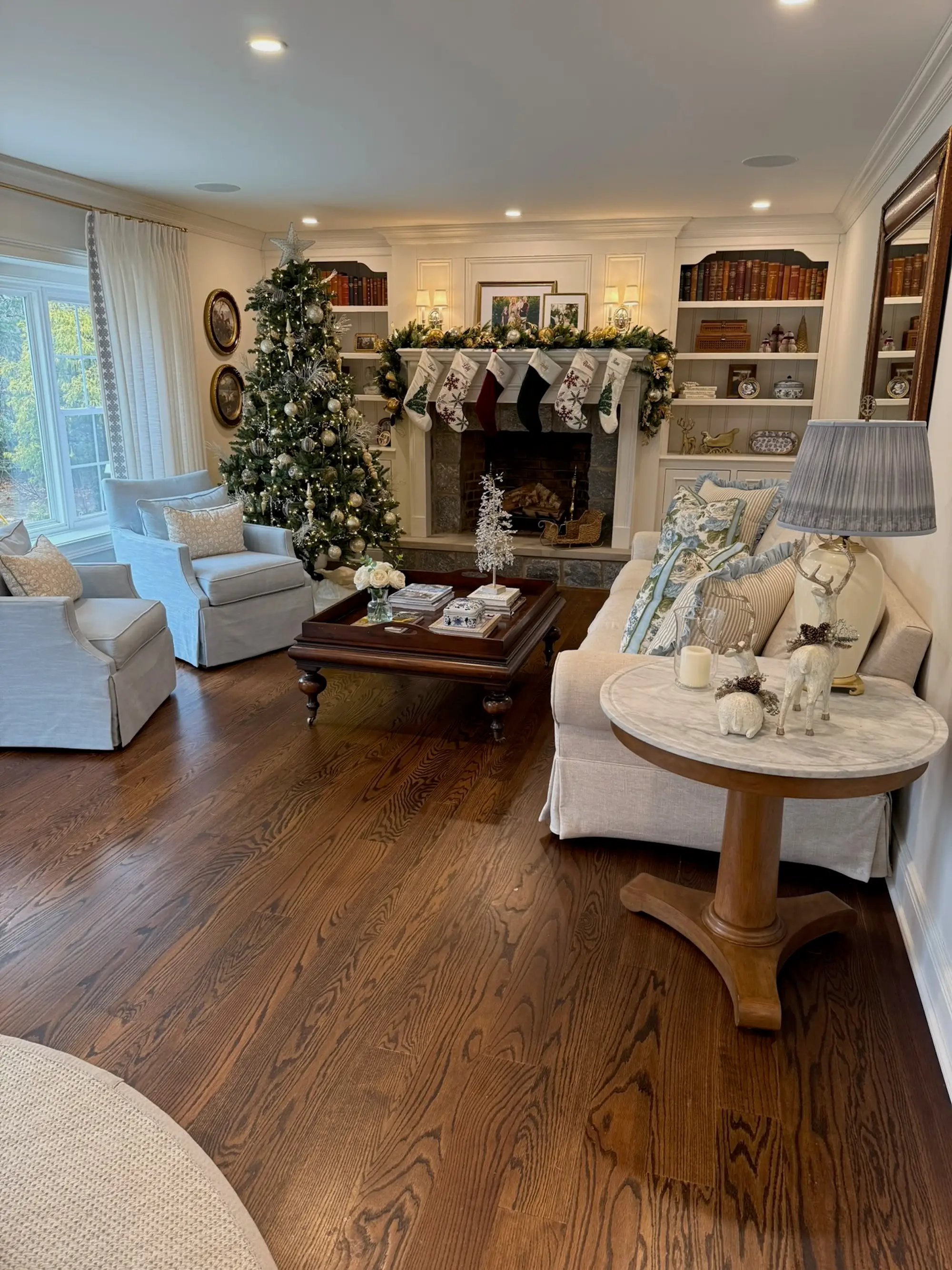 A living room with dark wooden floors, a white fireplace, and built-in bookshelves. The room is decorated for Christmas.