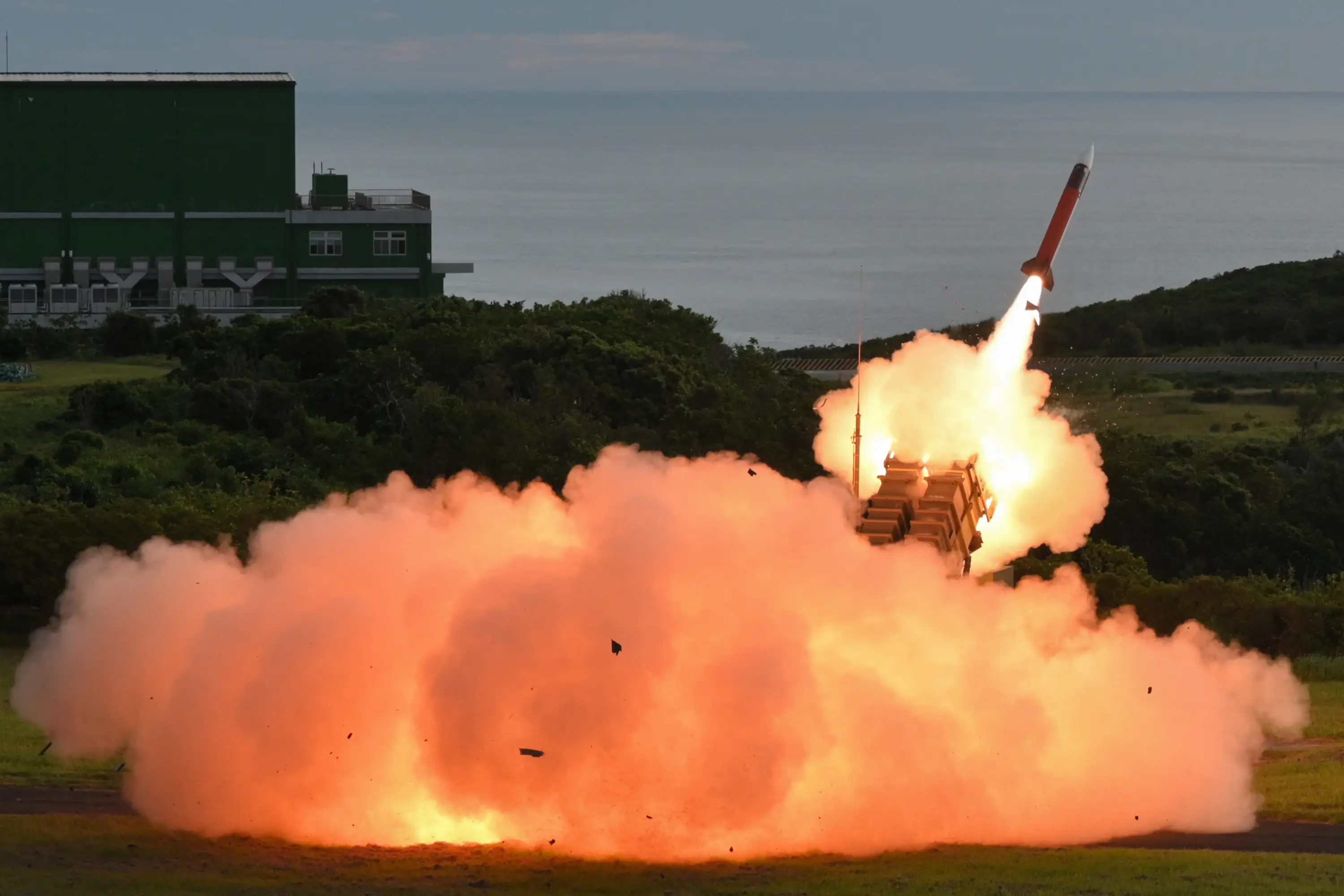 A red missile fires from a large piece of weaponry with orange smoke coming out of it, with fields and the sea in the background