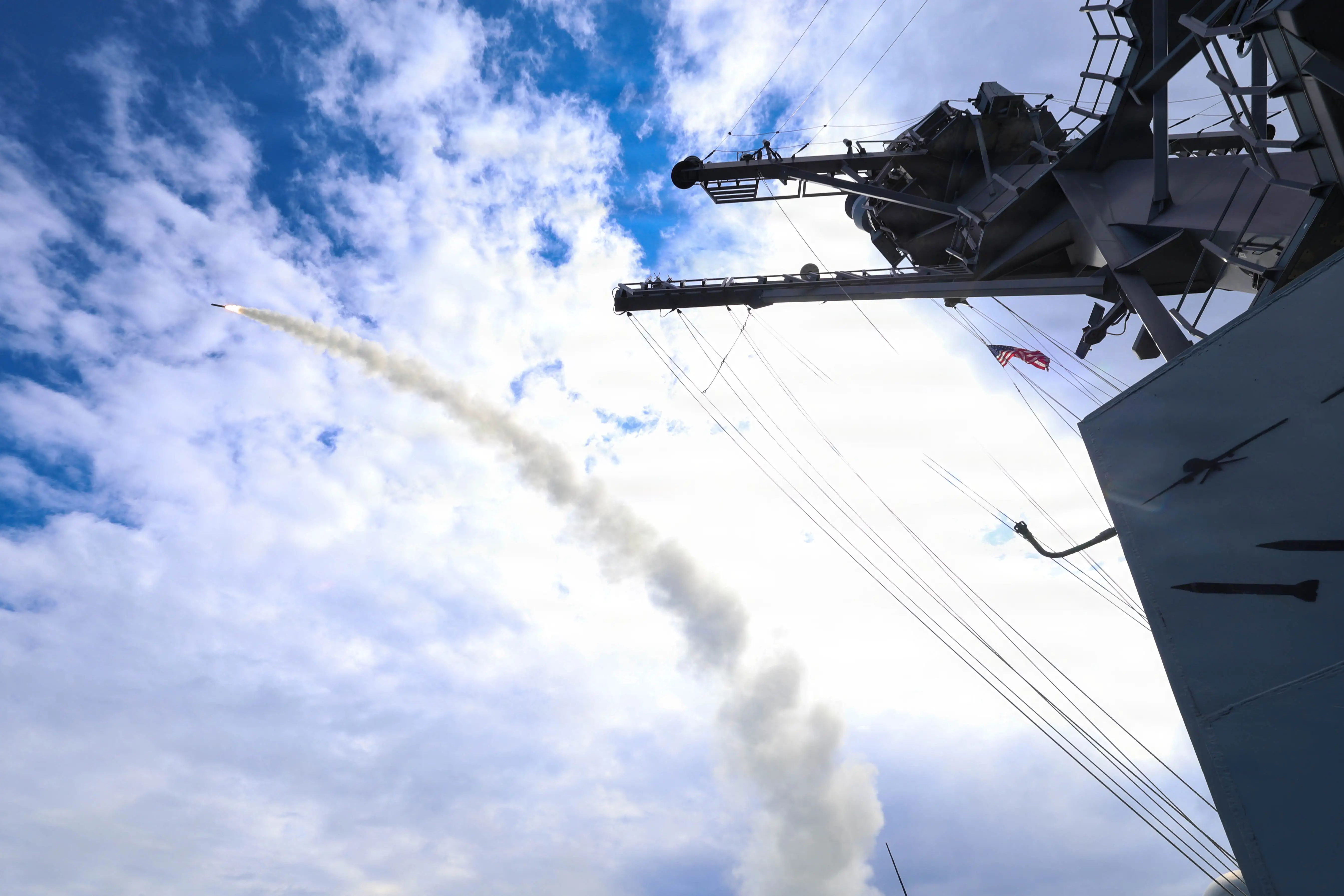 A missile arcs through a blue sky, with the gray structure of a US Navy ship imposing overhead from the right.
