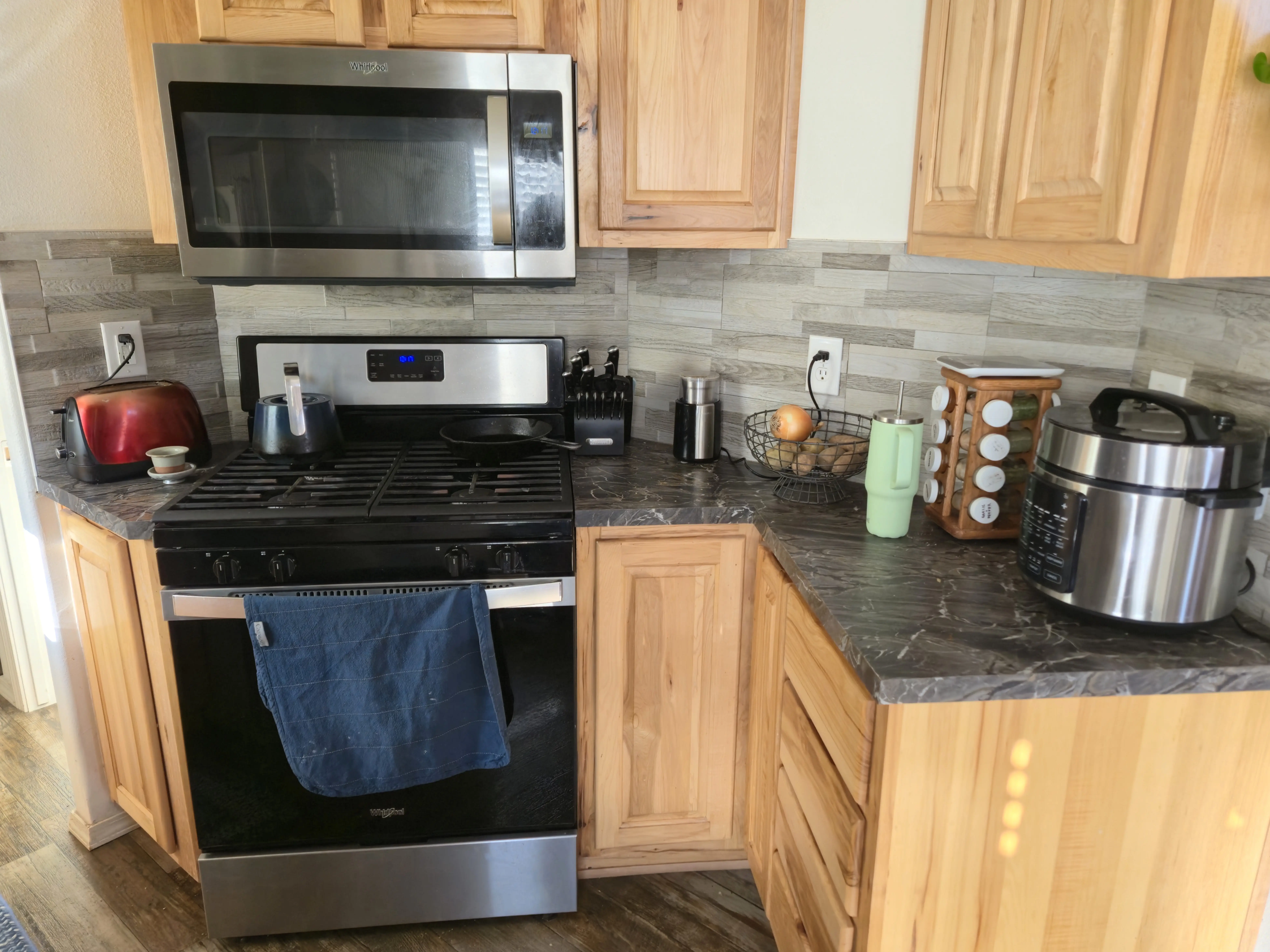 Light-wood kitchen cabinets with light gray backsplash, gray counters an stainless steel microwave above oven, rice cooker and spices on counter