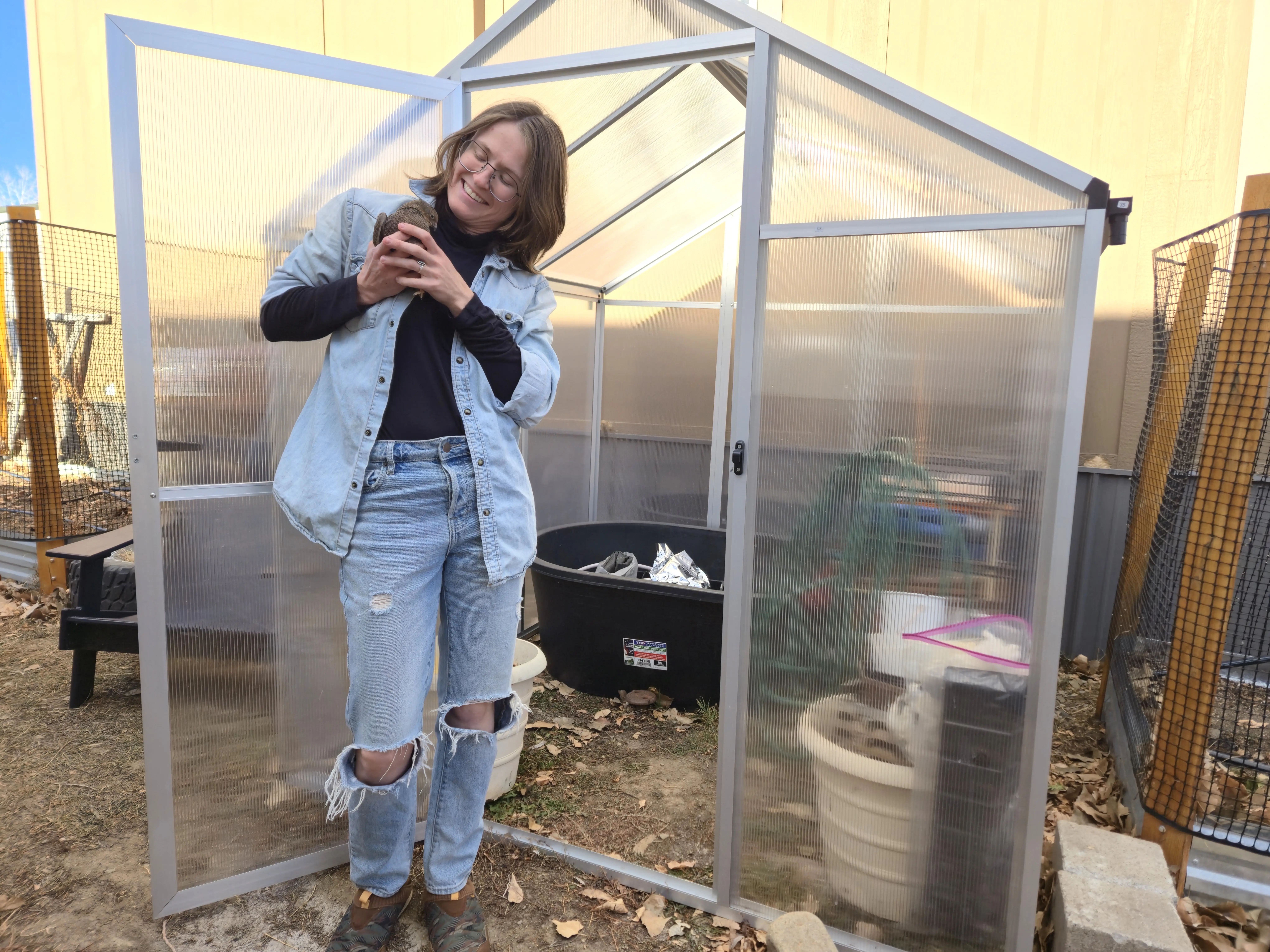 Author Amber McDaniel smiling, holding quail next to outdoor greenhouse