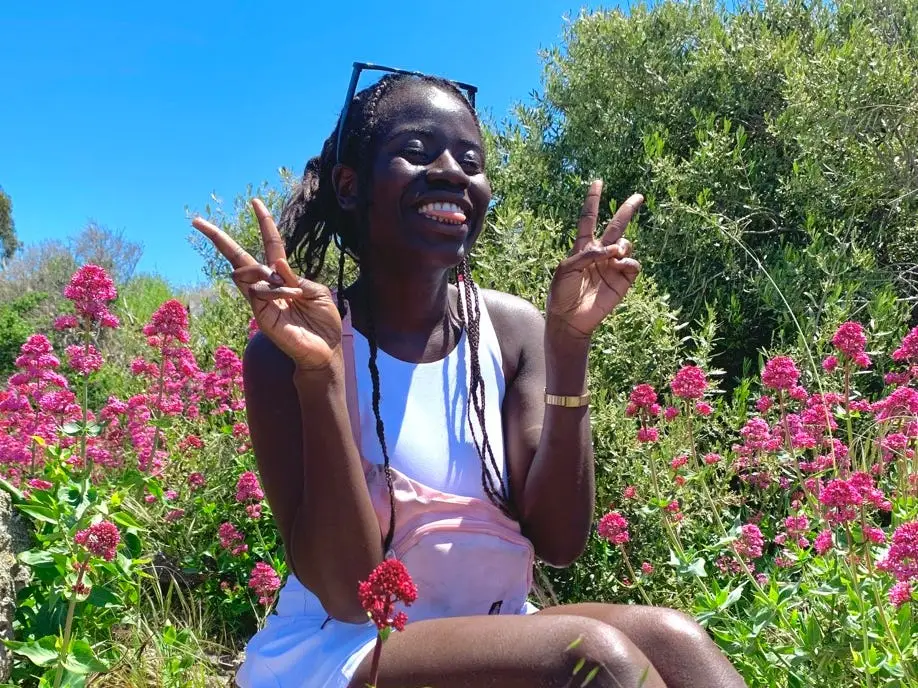 Author Alicia Simba smiling while sitting, holding up peace signs, in front of flowers