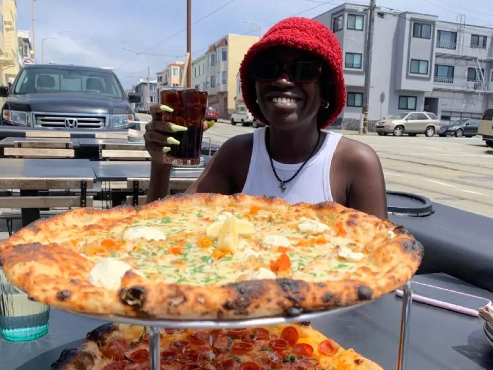 Author Alicia Simba smiling with a beverage in hand and pizzas in front of her