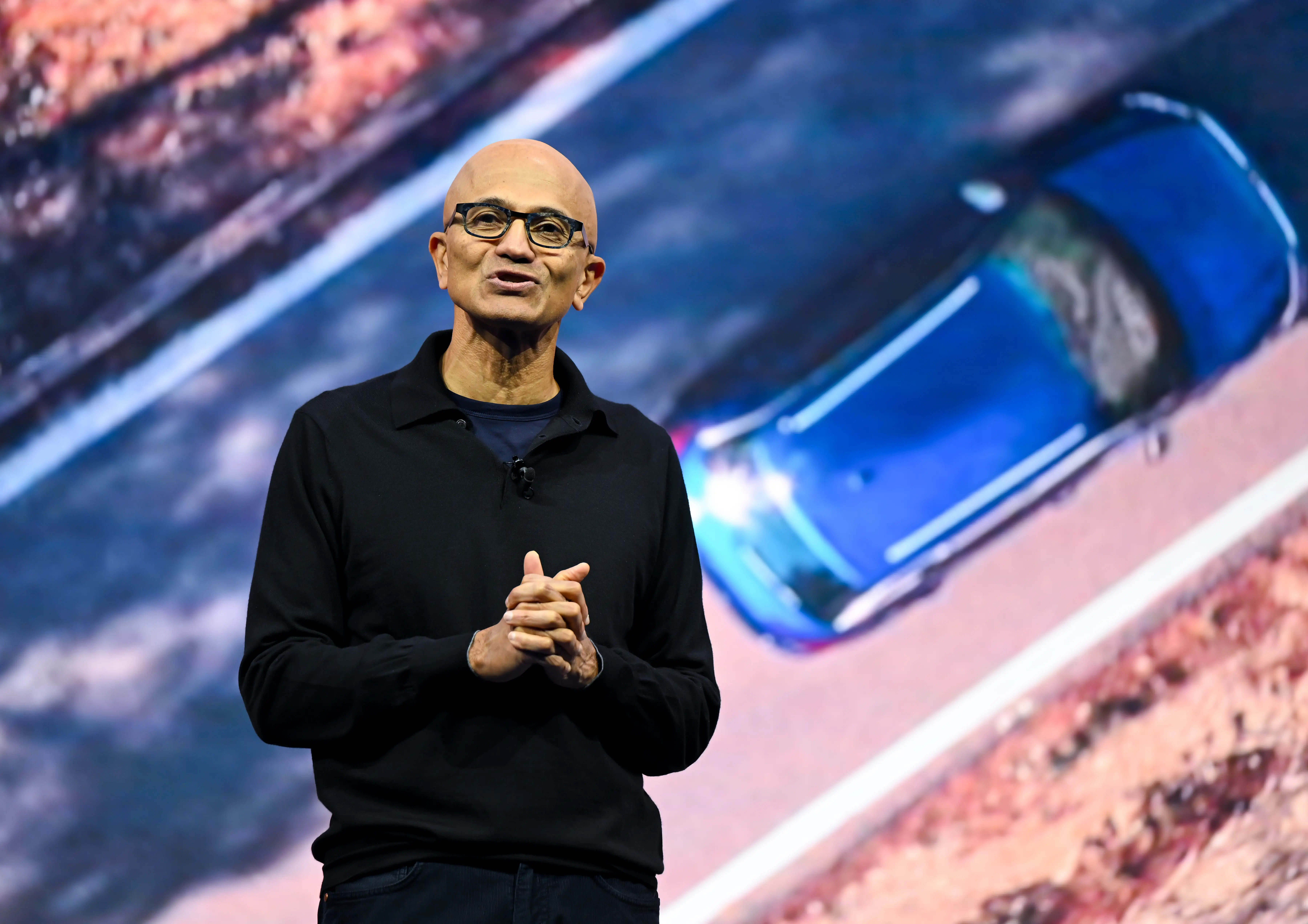 Satya Nadella, dressed in a long-sleeve black polo shirt, clasps his hands as he speaks onstage. The screen behind him shows a bird's-eye view of a blue car driving down a road.