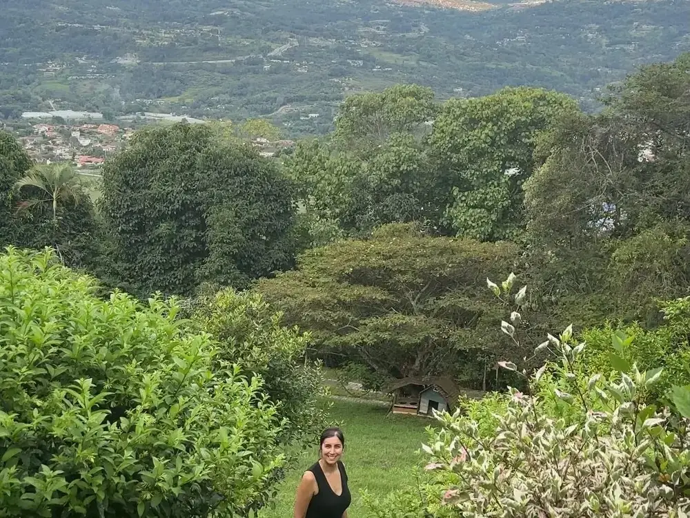 A woman stands on a hillside surrounded by trees.