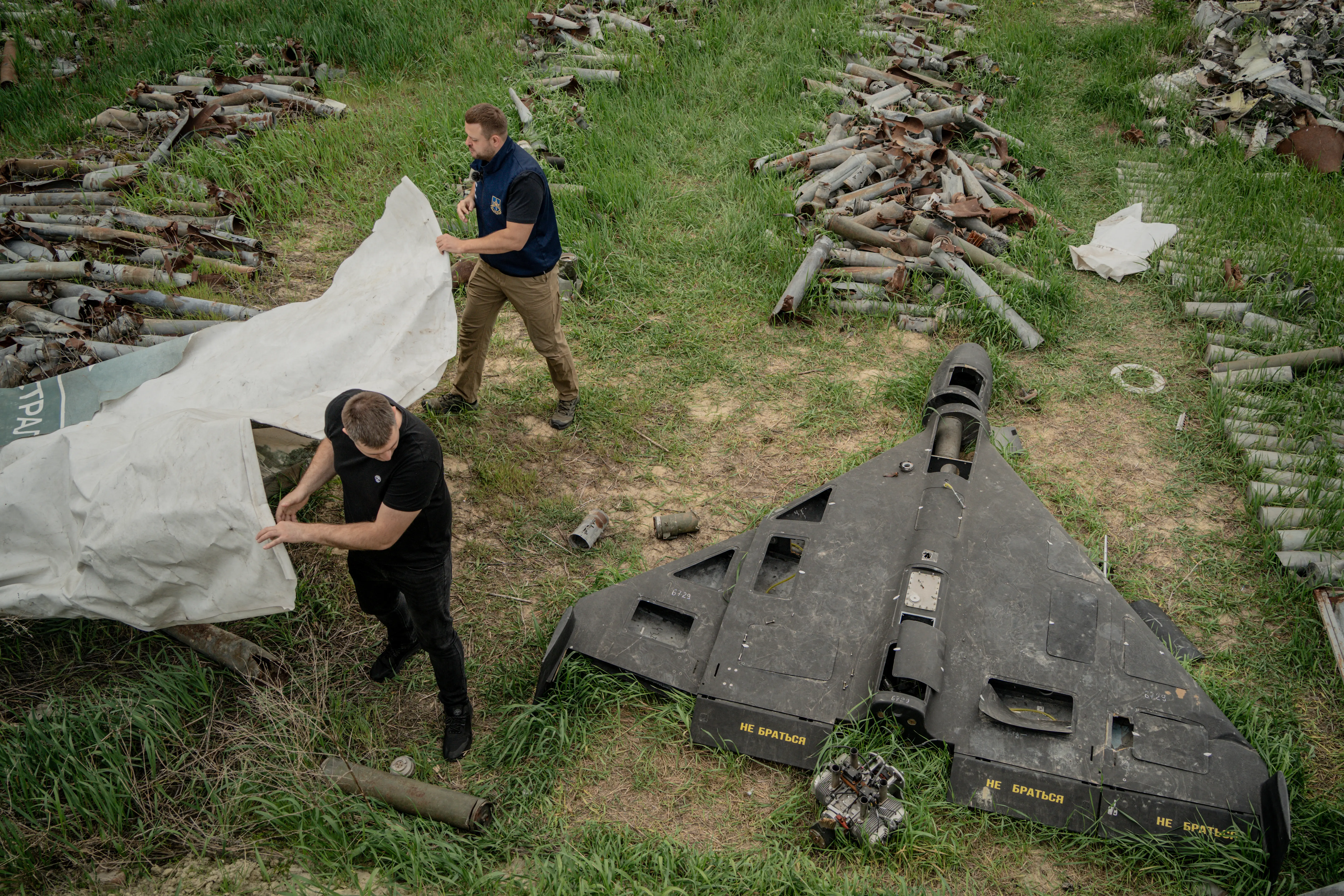 A damaged large black drone and other scraps lying in a field with two men standing beside holding a white tarp