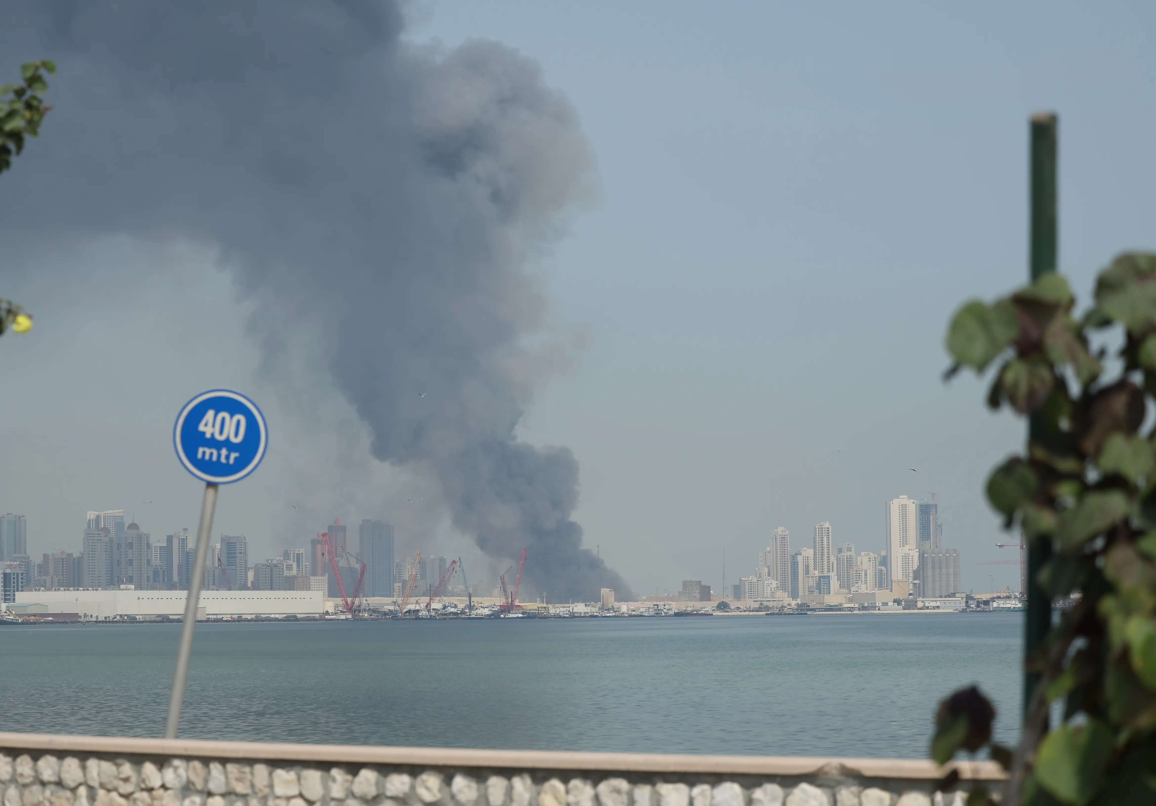 Smoke rises from a skyline with water in the foreground under a blue sky