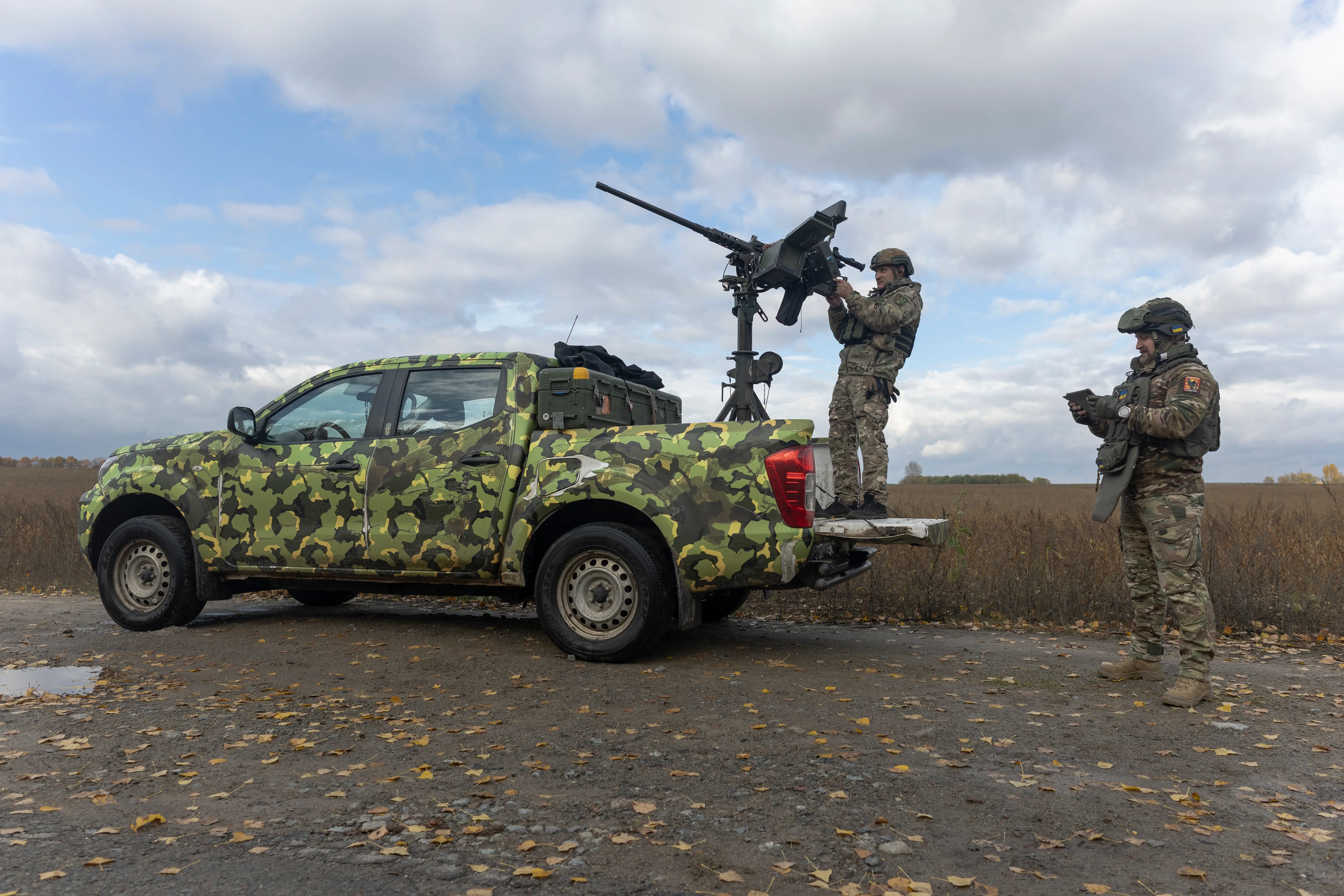 A man wearing camoufage stands on the back of a camoufage-painted truck pointing a weapon into a cloudy and blue sky, with another man standing beside