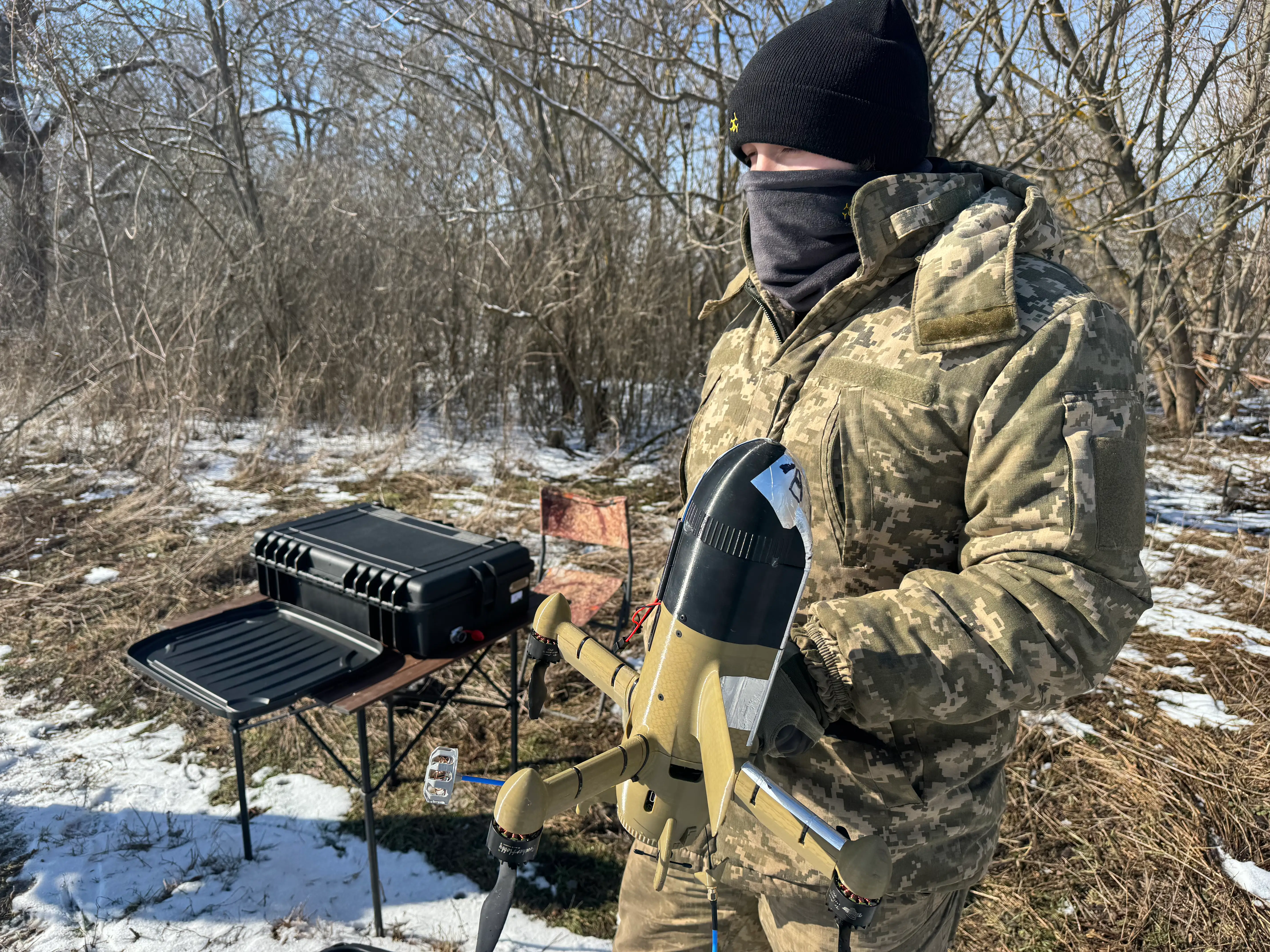 A man in camouflage gear and a black beanie stands in a snowy field in front of trees holding a black and beige drone, standing beside black equipment