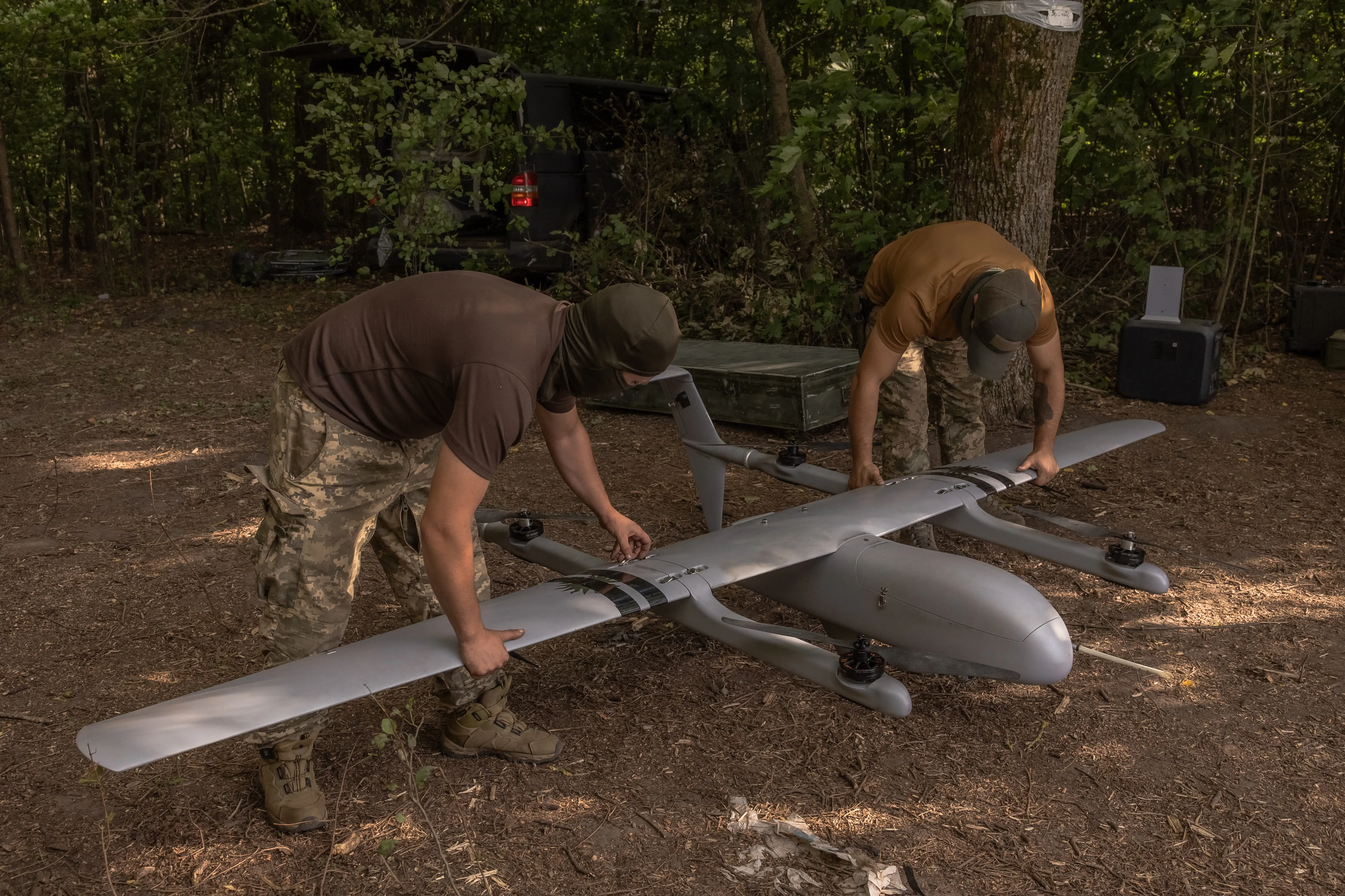 Two men bending over holding a large grey drone between trees