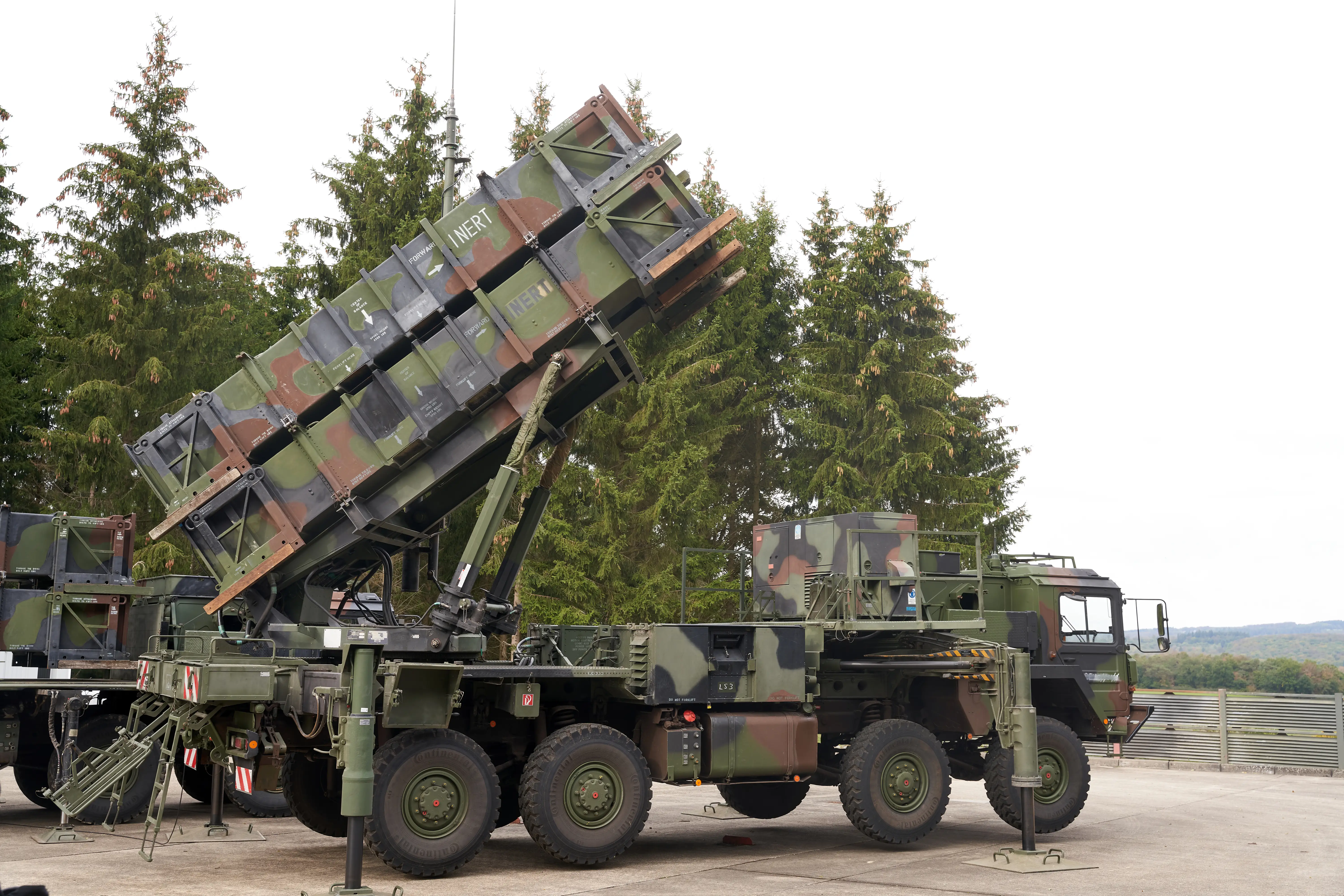 A large camouflaged truck-mounted weapon beside trees and under a white sky