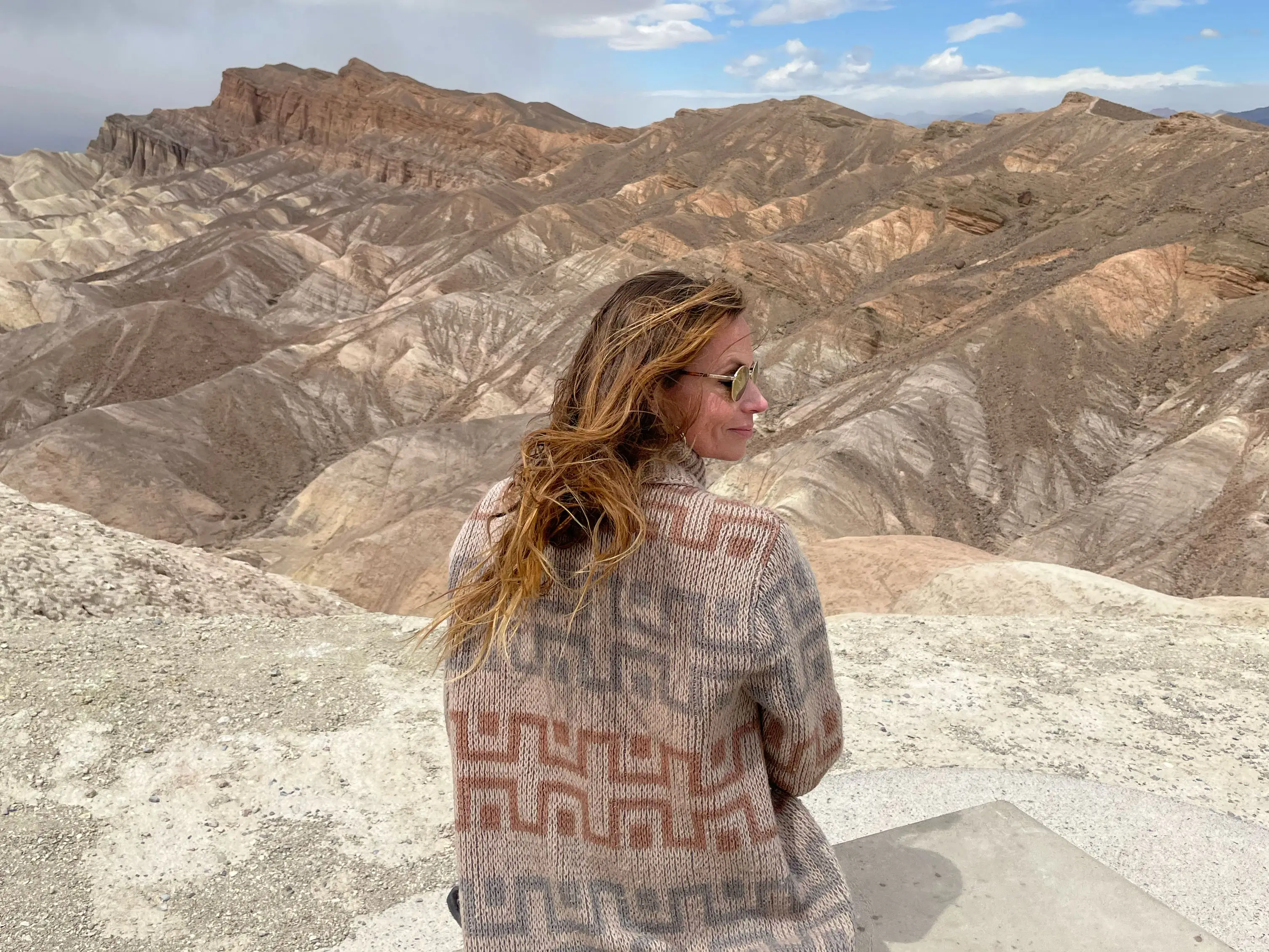 Emily sits on a mountain, overlooking layered desert hills under a partly cloudy sky.