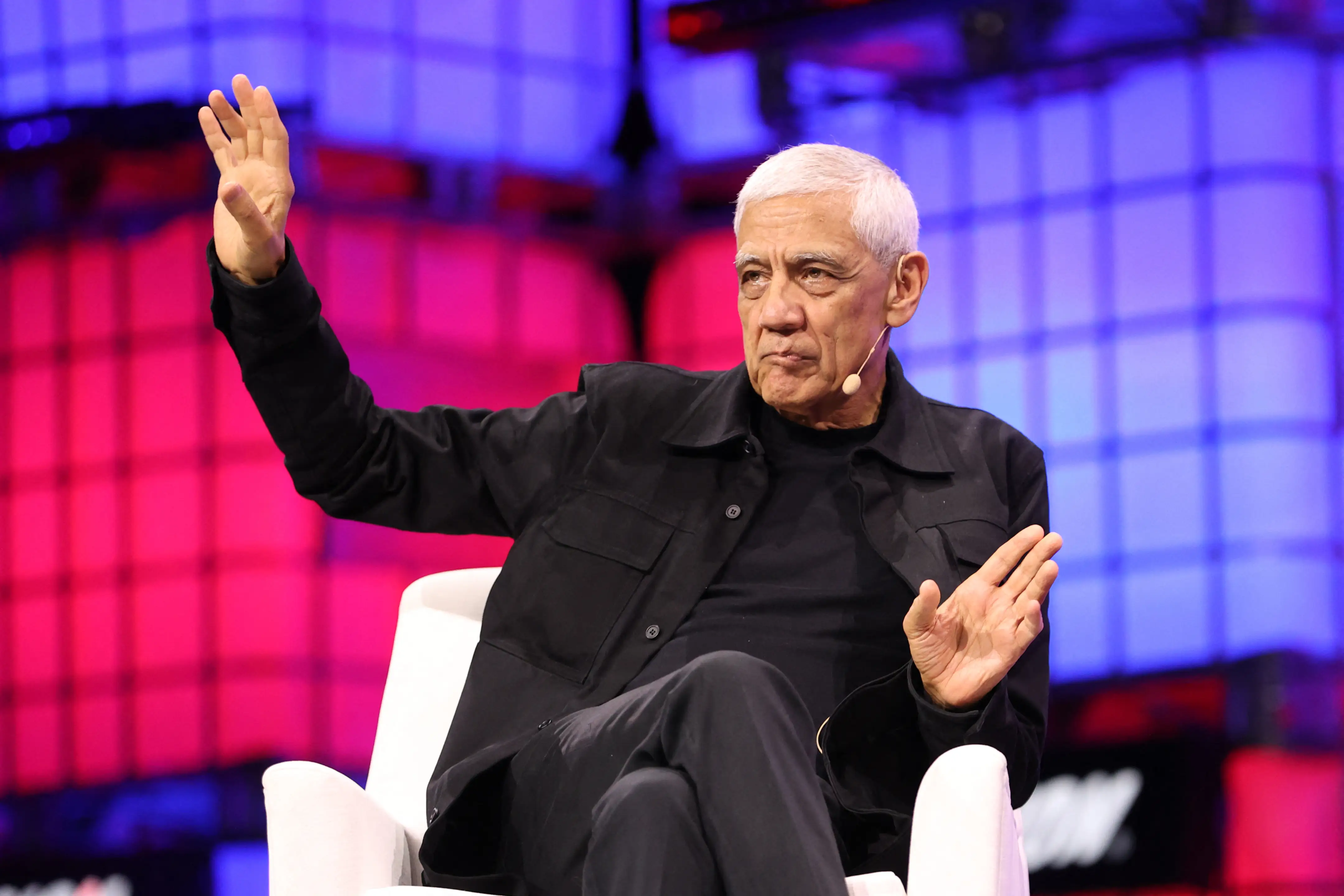 Vinod Khosla, wearing a black button-up jacket over a black T-shirt. He is on stage, sitting in a white chair, with his right hand raised.