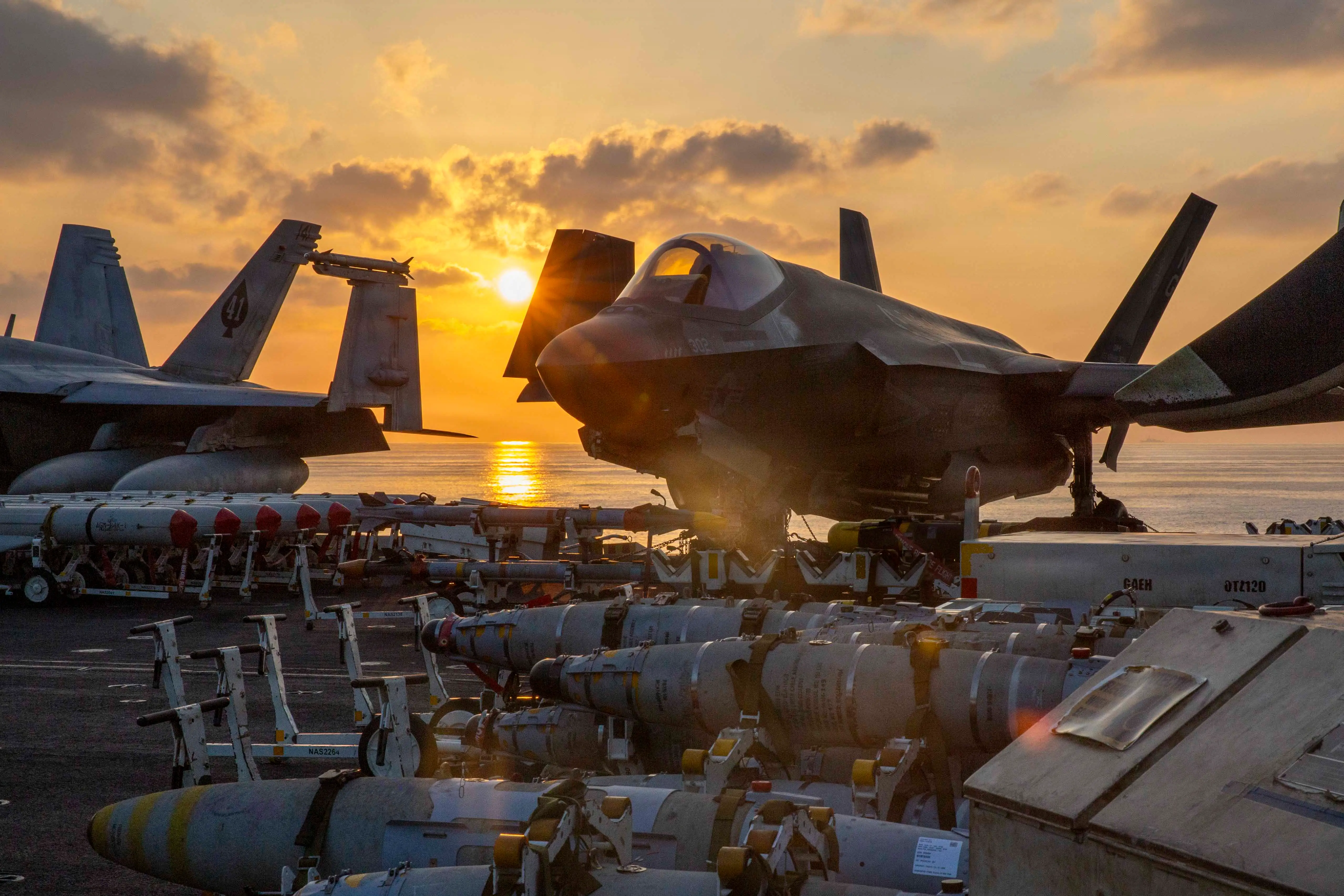 Two F-35 aircraft sit on an aircraft carrier surrounded by munitions. The sun is setting in the background.