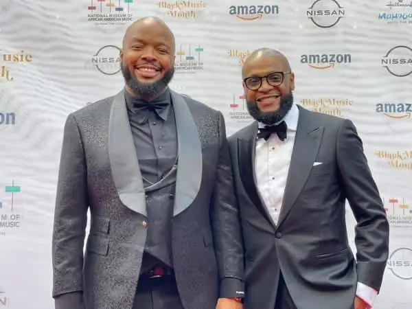 Two men dressed in tuxedos standing in front of a white backdrop