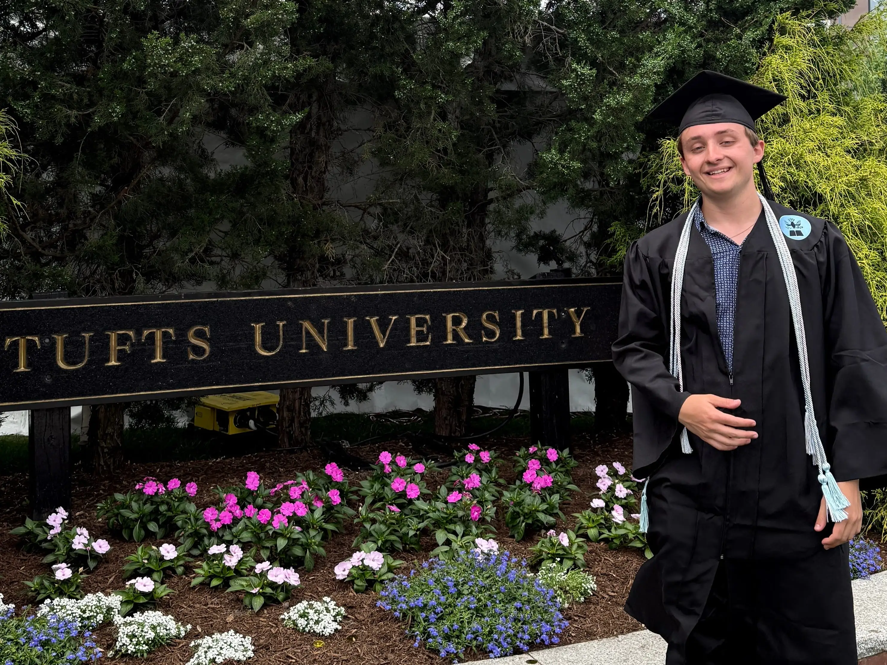 Oliver Fox in front of the tufts university sign wearing a graduation gown