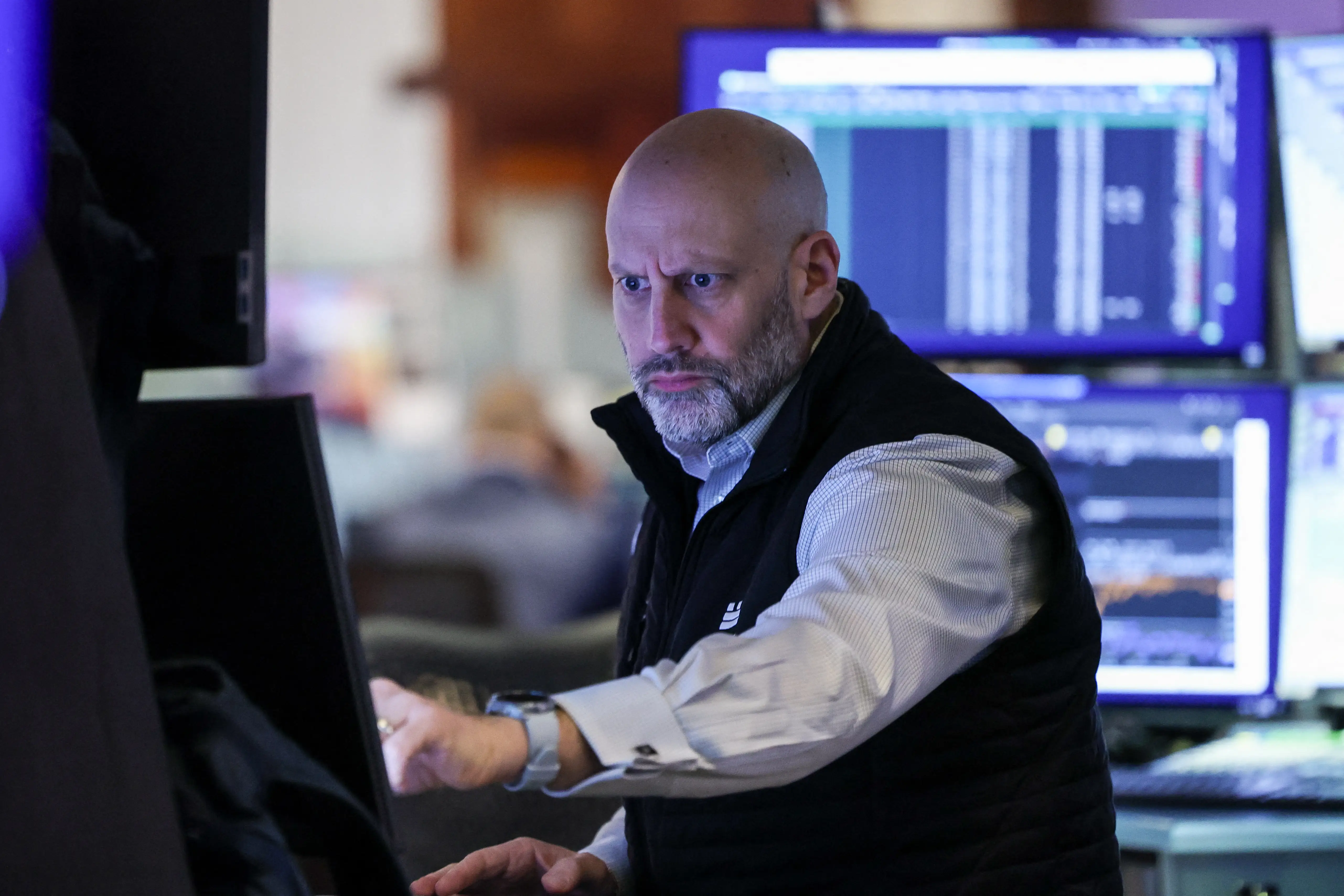 A Wall Street trader, dressed in a long-sleeve button-up and a black vest, points at a computer screen. His face is scowling.
