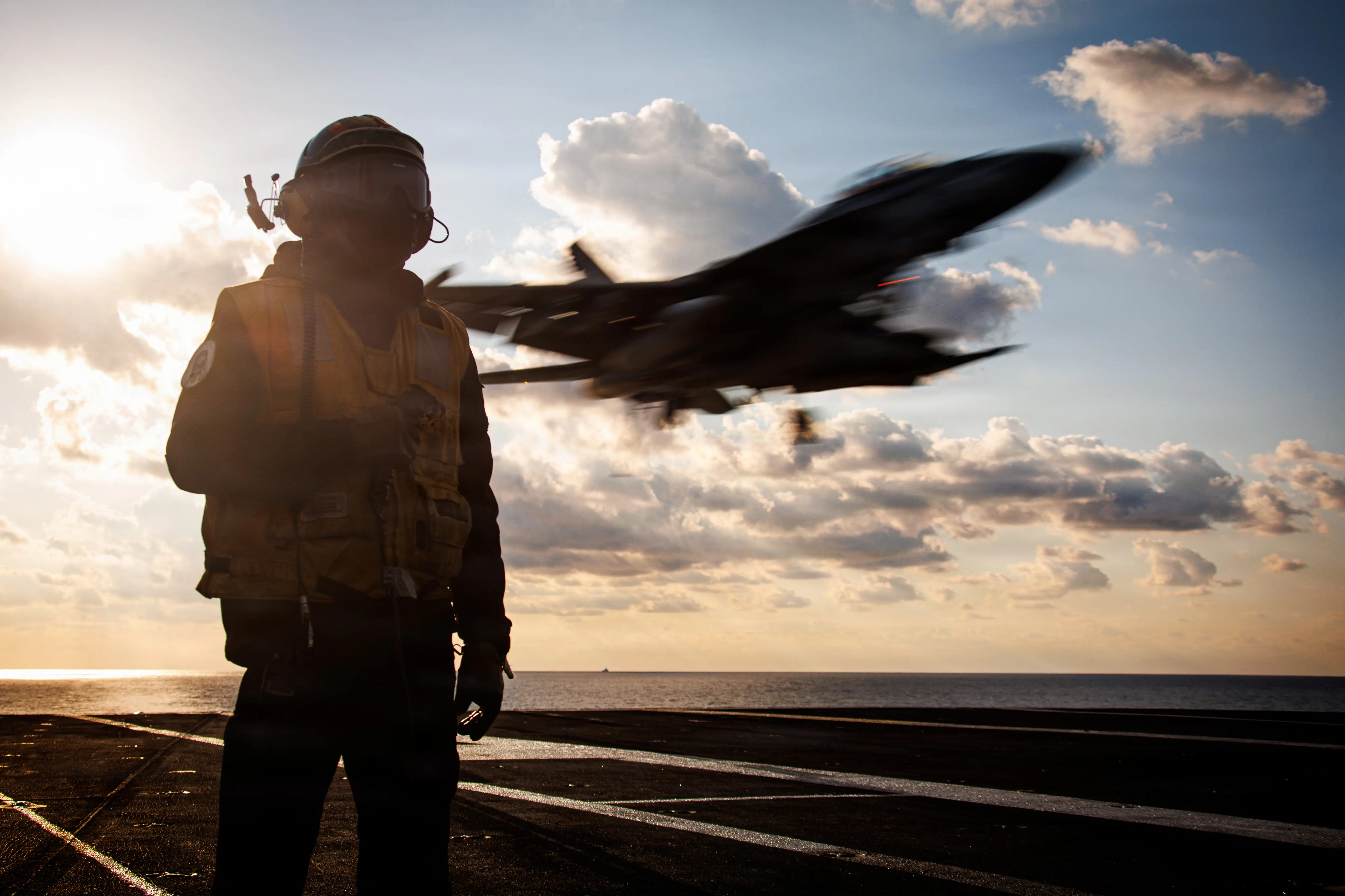 A man stands on the deck of an aircraft carrier as an F-35 flies by.