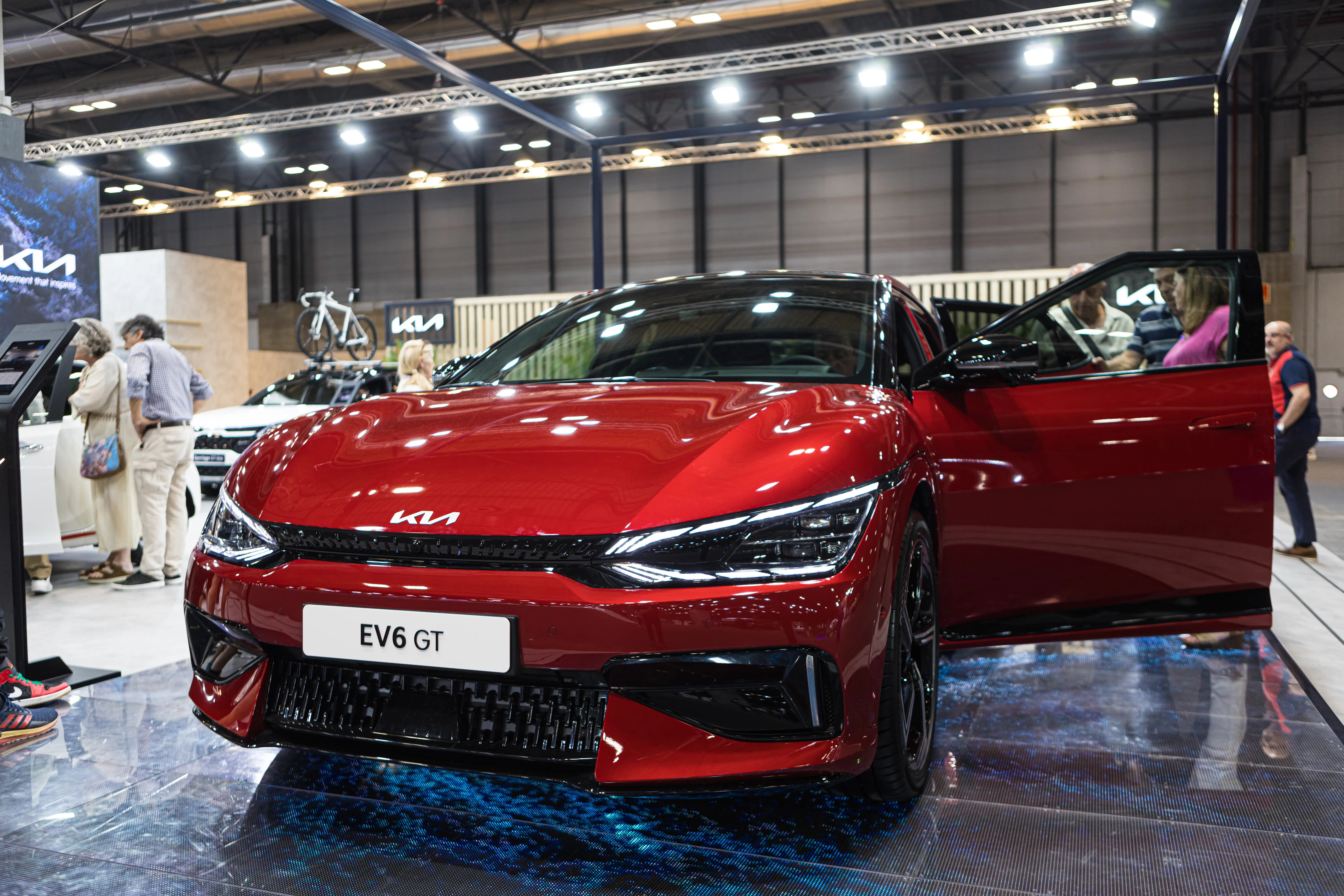 A red Kia EV6 GT is parked on a showroom floor with the headlights on, and the driver's side doors open.