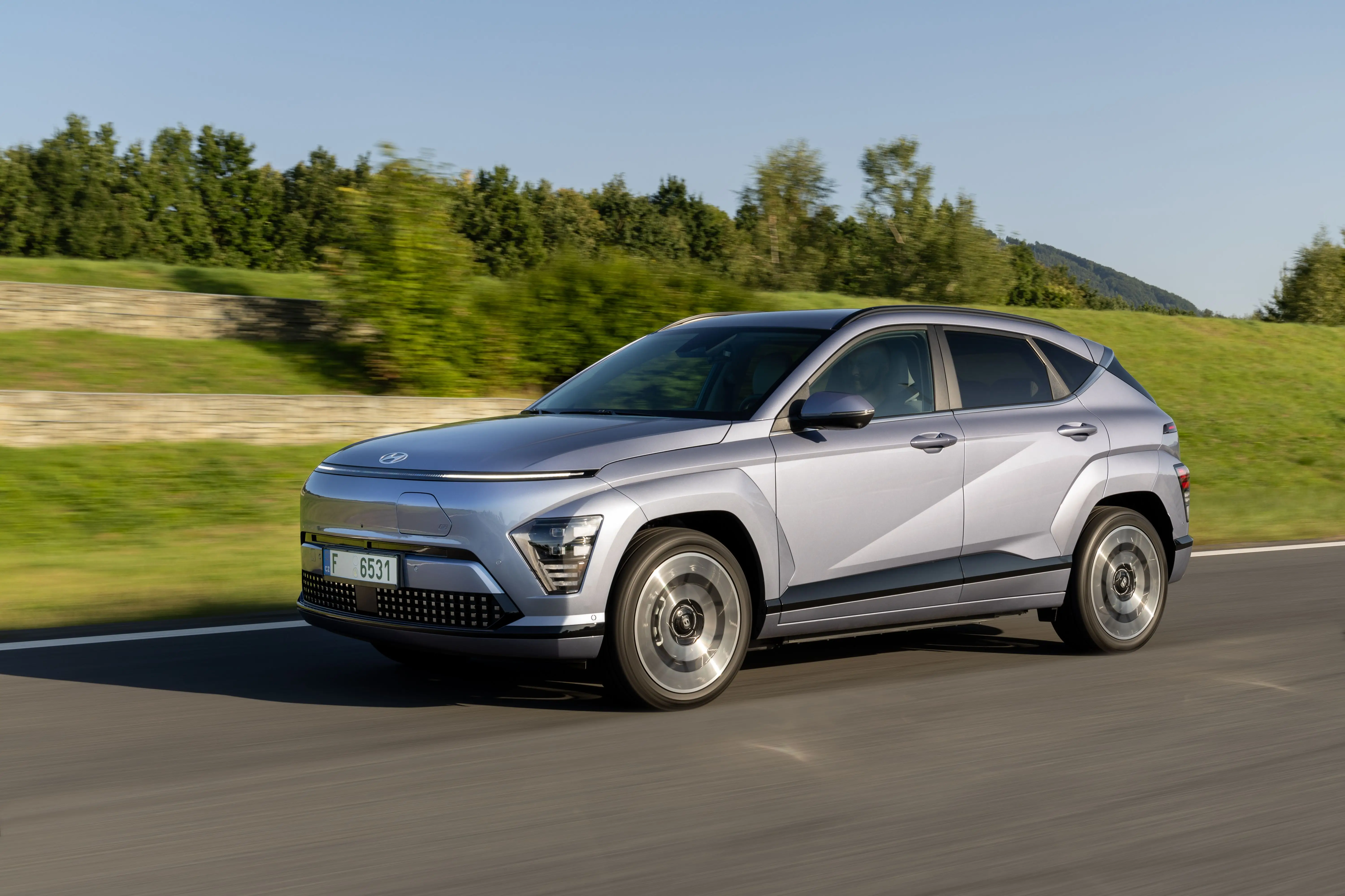 A silver Hyundai Kona electric is driving down a roadway. It's passing a grassy field and some blurry trees.