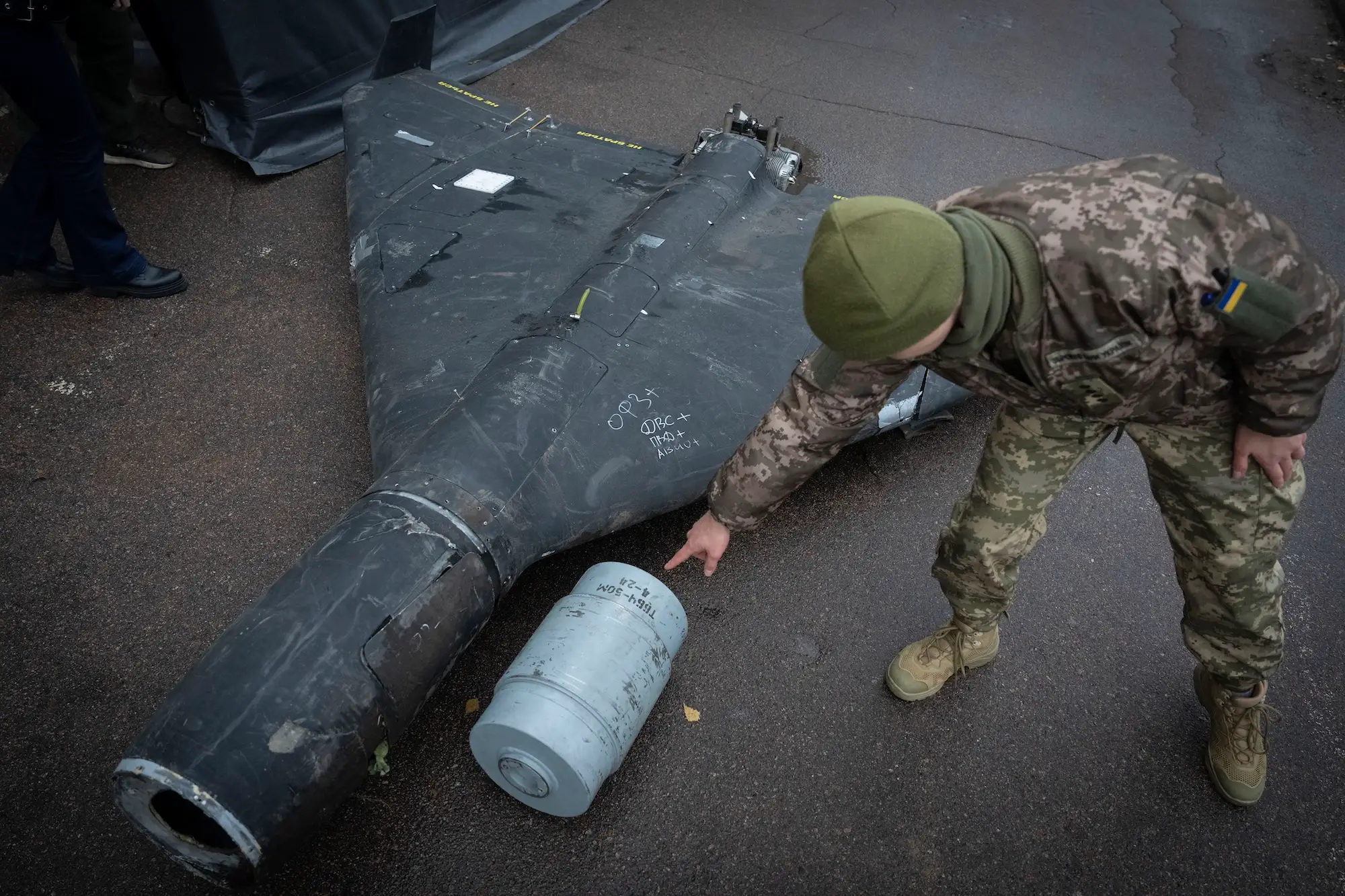 A large black drone on the ground, with a man in camouflage bending beside it and gesturing to a small metal drum-shaped object beside it