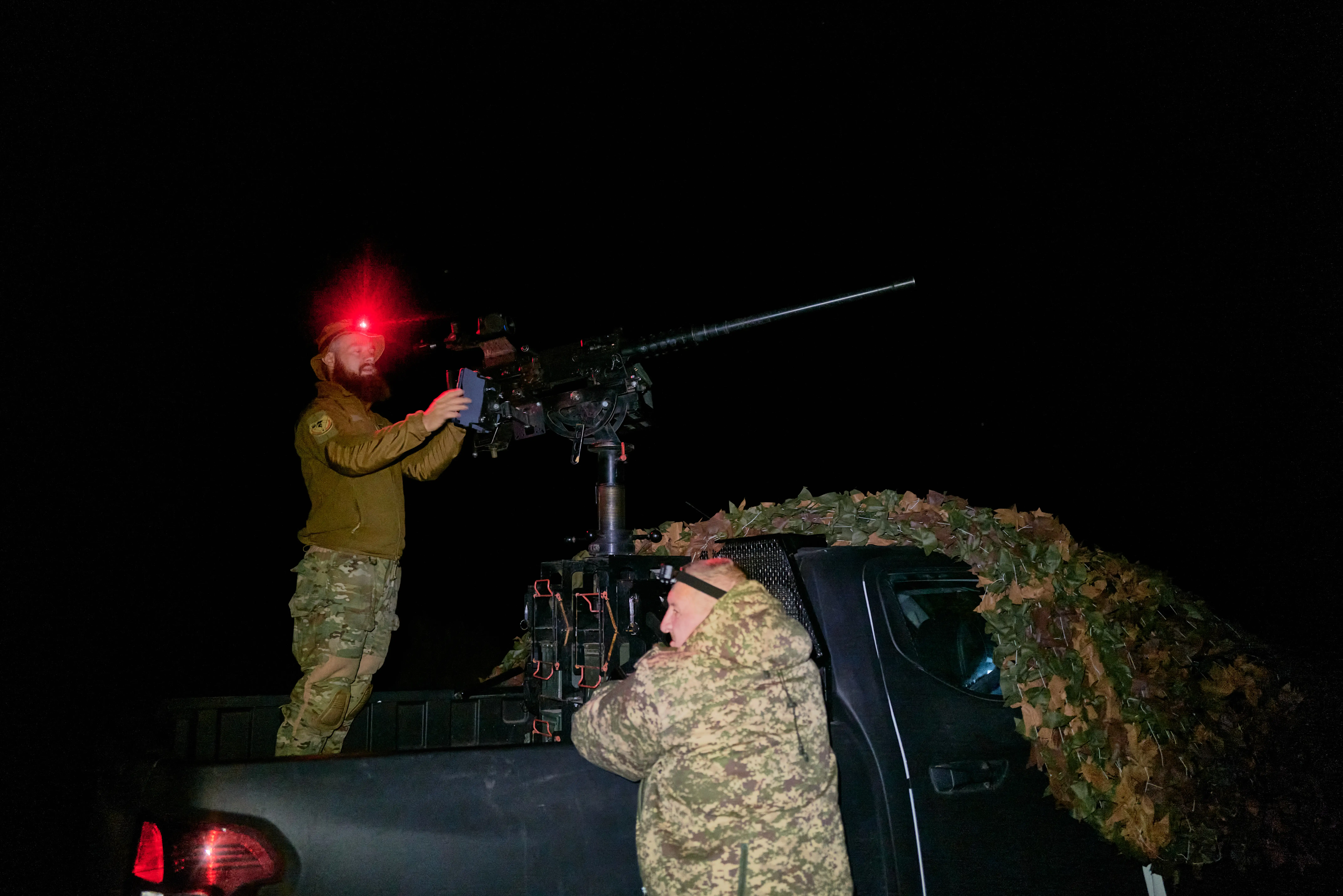 A man in camouflage stands on the back of a black truck pointing weaponry in the air, with another man in camouflage standing beside it, all under a black sky