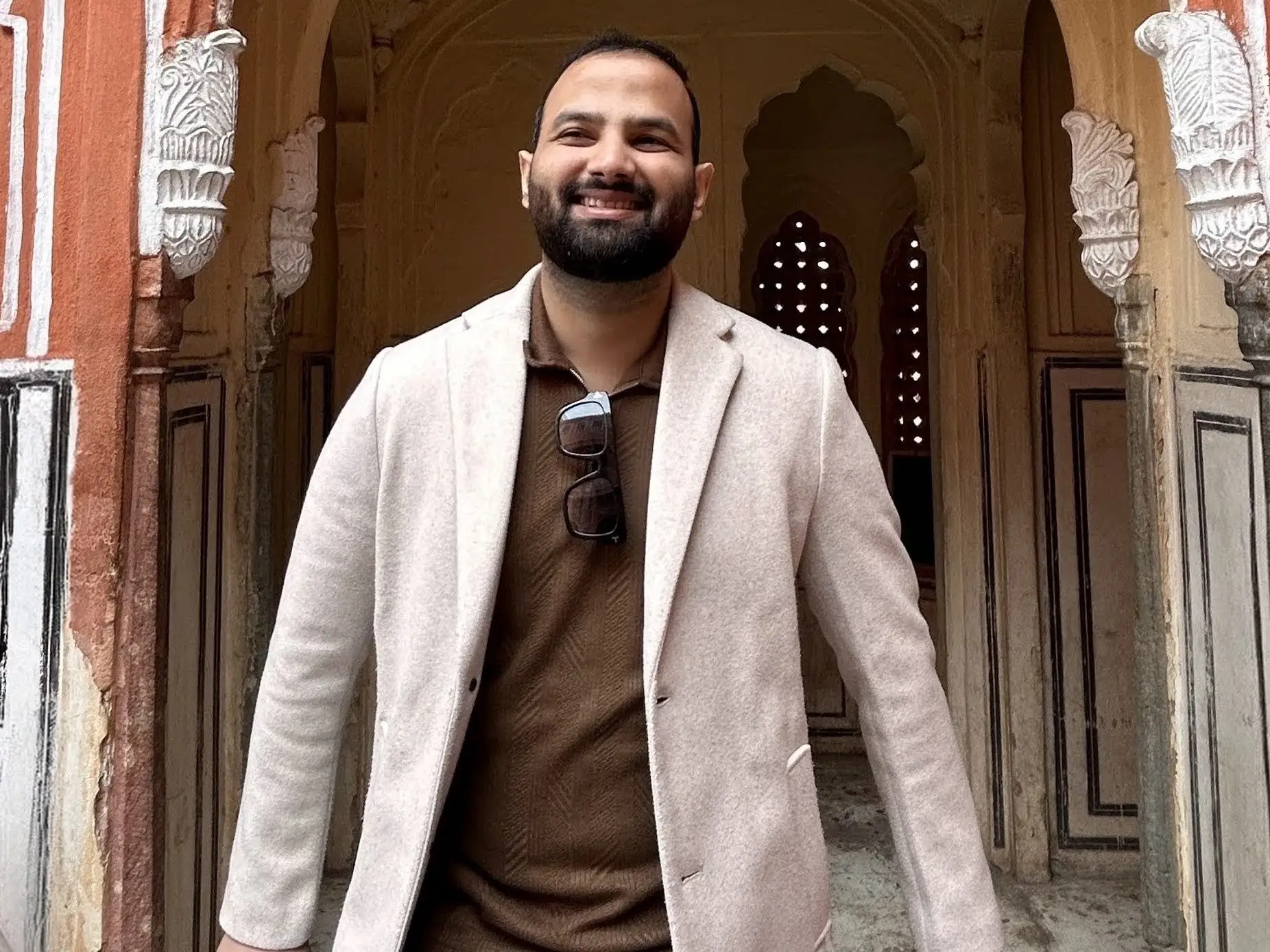 Smiling, a bearded man in a beige coat stands beneath an ornate red and white arched doorway with carved details.