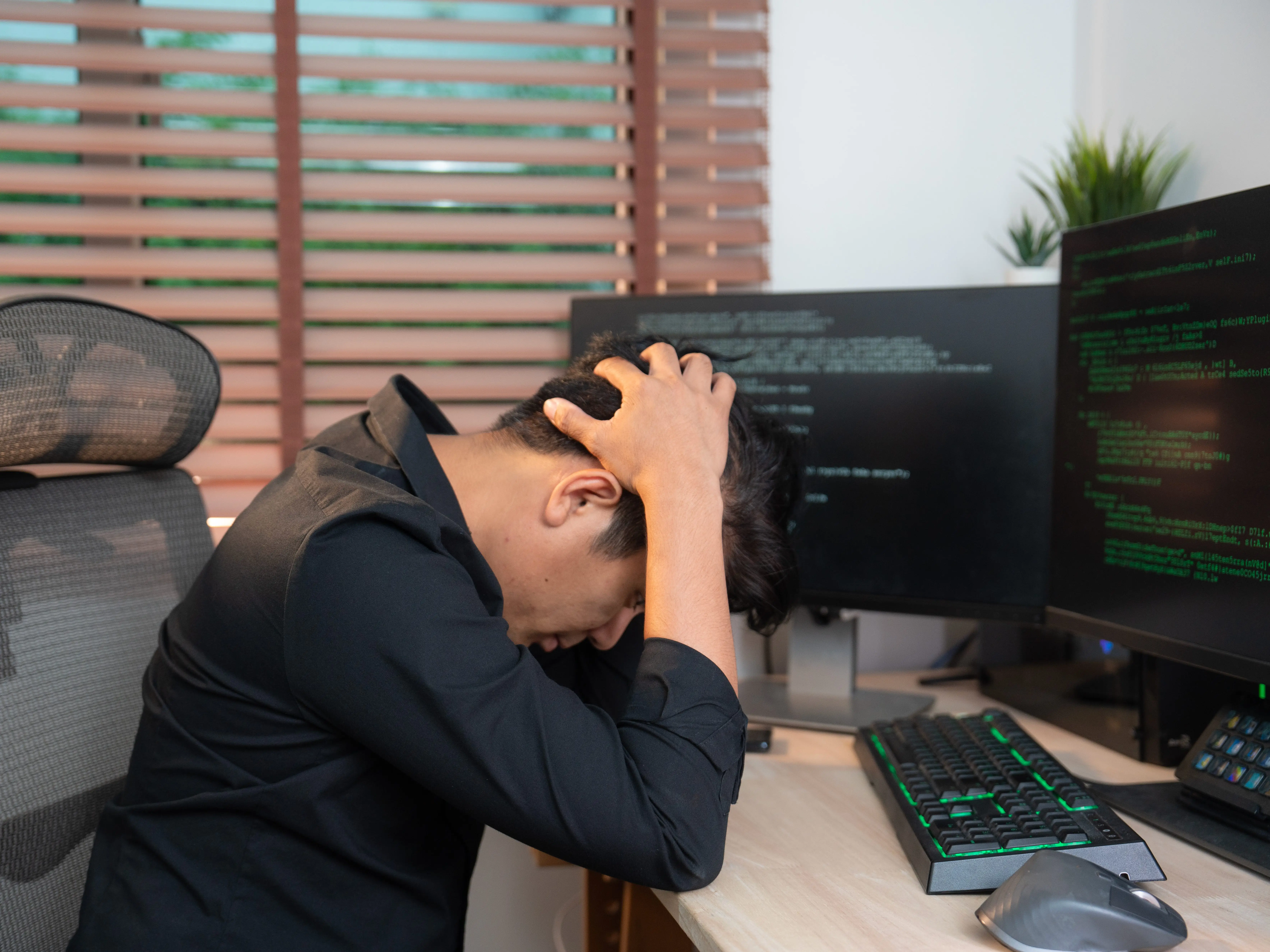 A person wearing a black shirt has their hands on their head while sitting at a desk, and there are lines of code on the computer monitors in front of them