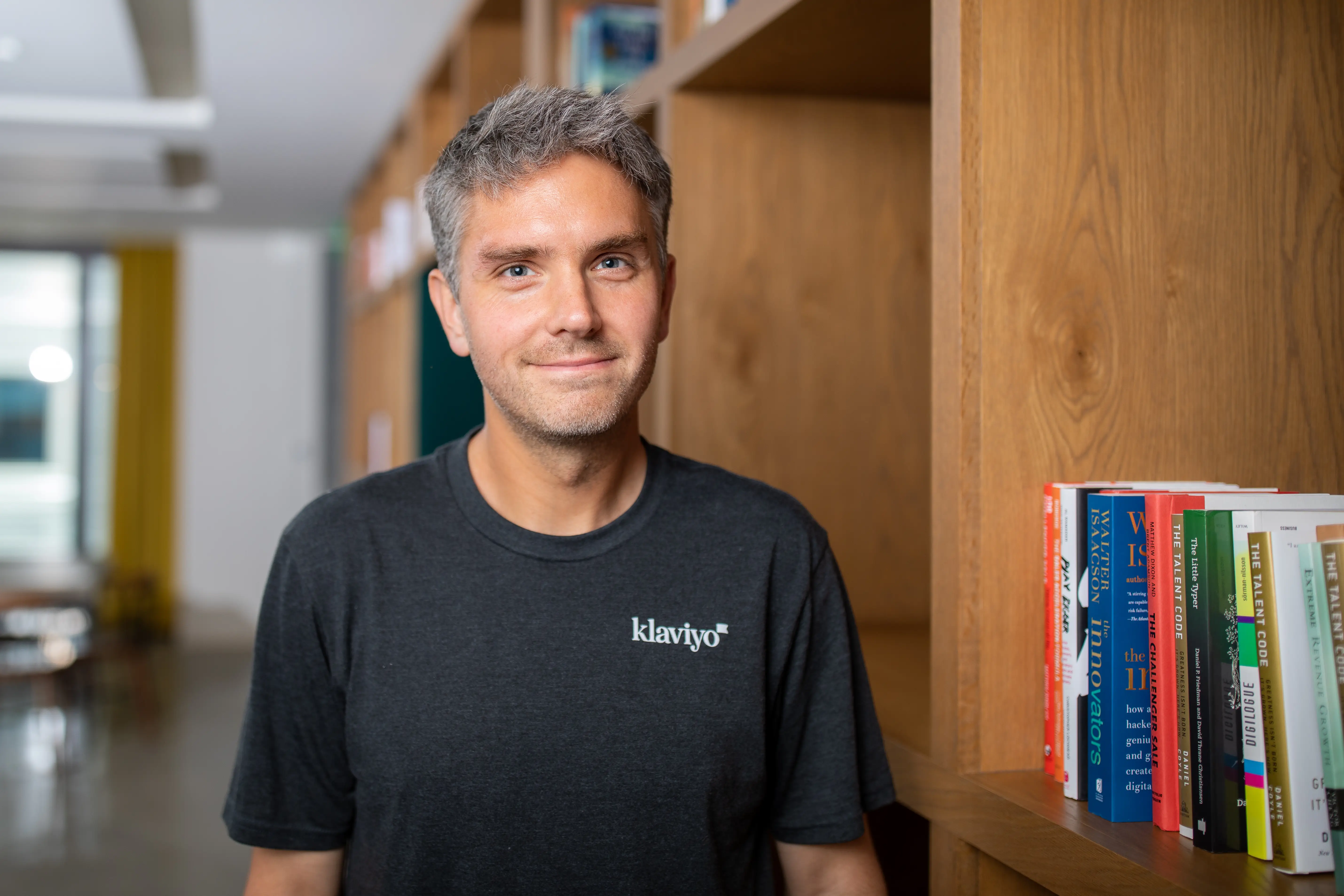 headshot of a man in a gray shirt next to a bookcase