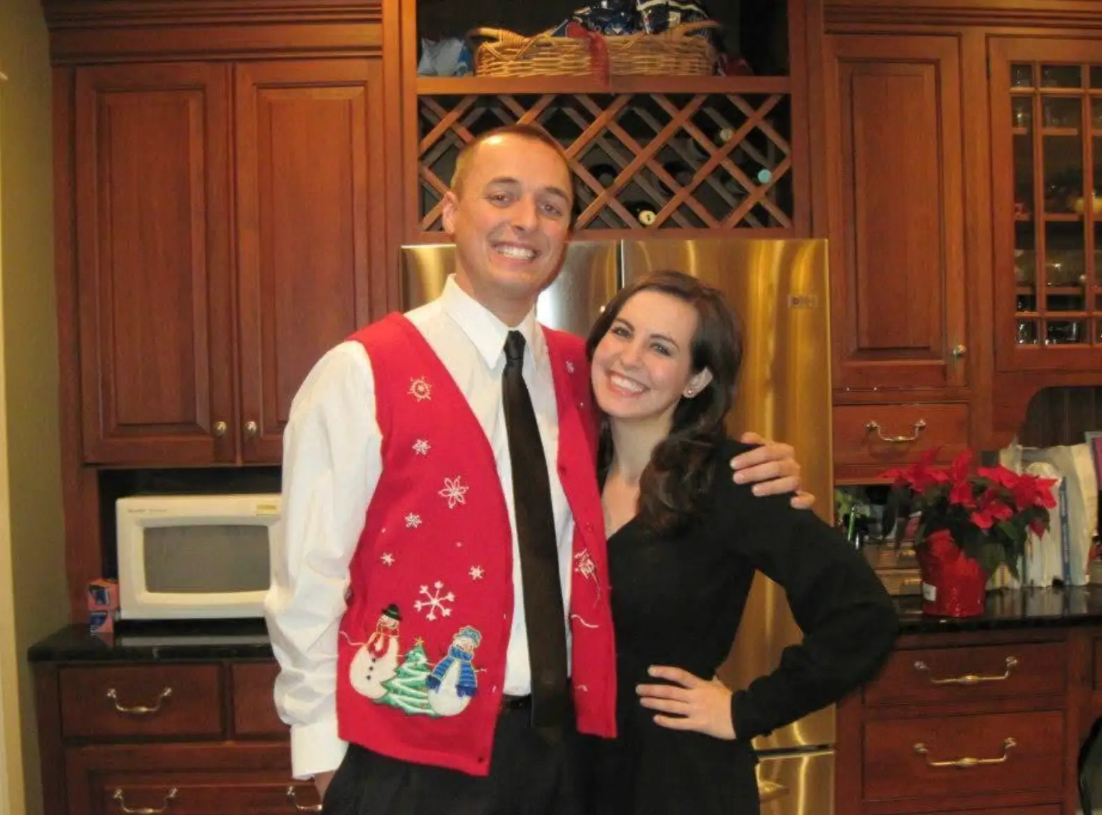The writer, wearing a black dress, and her husband, wearing a festive holiday vest, standing in their kitchen.