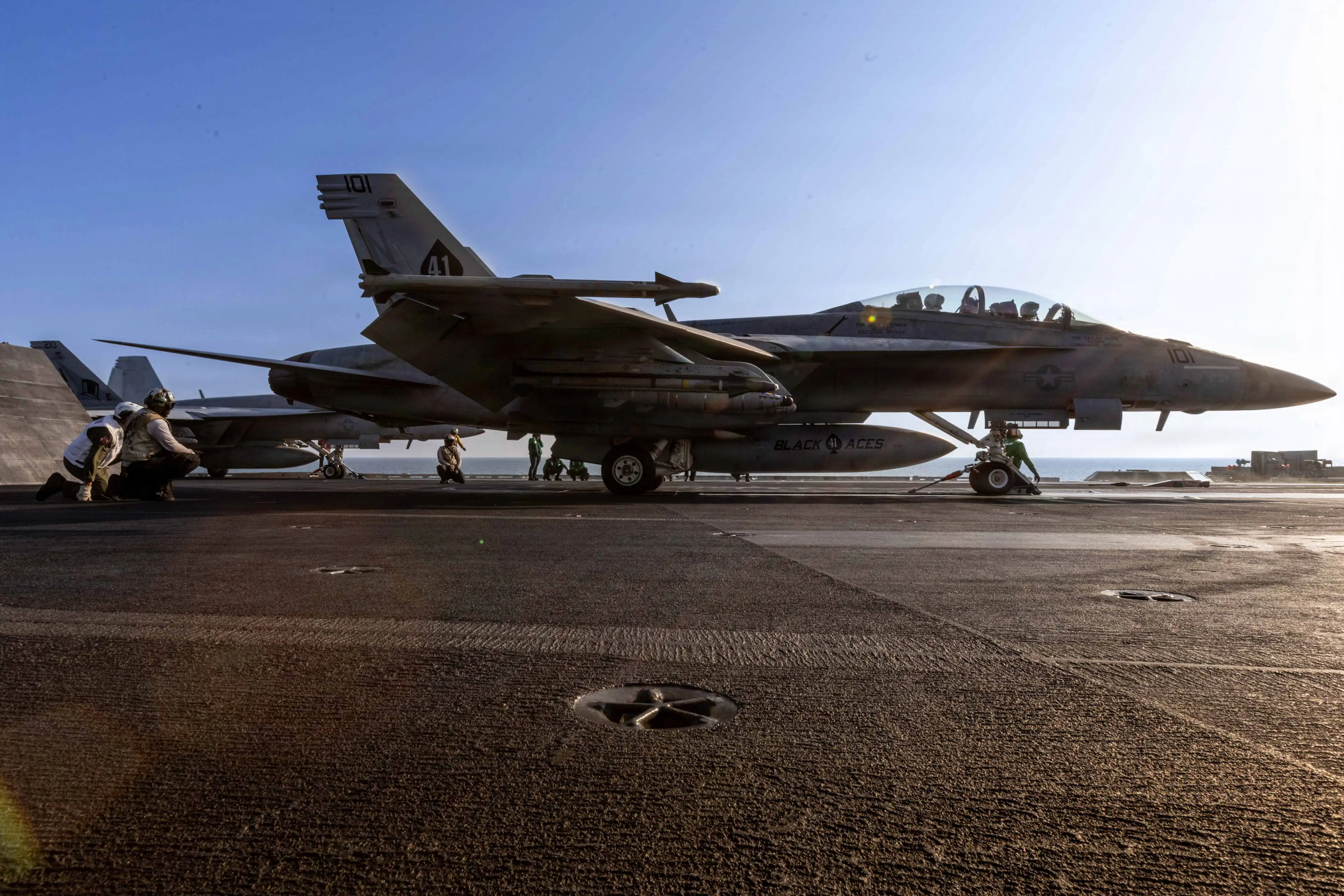 A US Air Force F/A-18 Super Hornet sits on an aircraft carrier with crew standing and crouching around it.