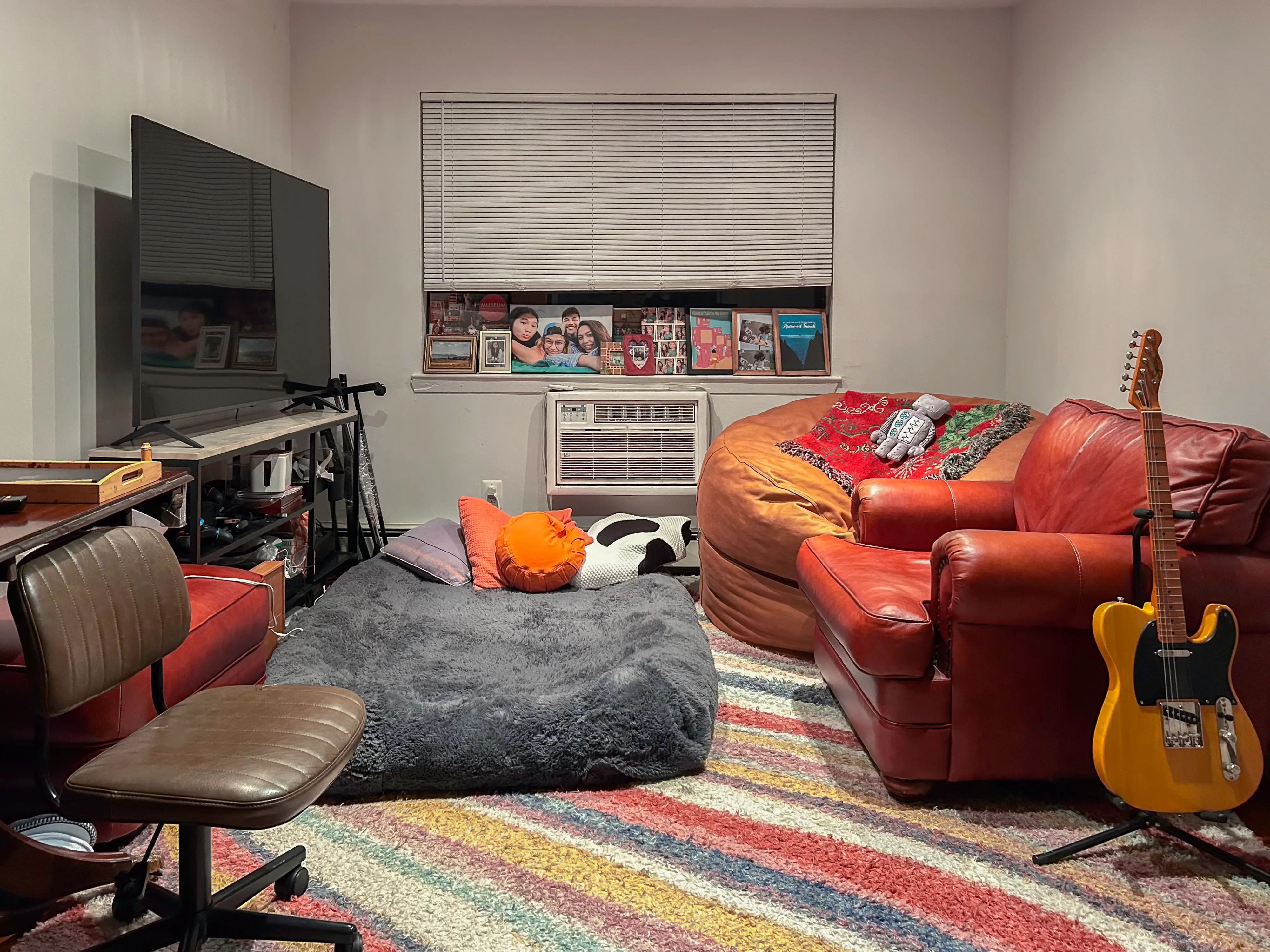 Inside a living room with a vibrant rug and seating areas, a TV on the left, a guitar on the right, and a window in the back