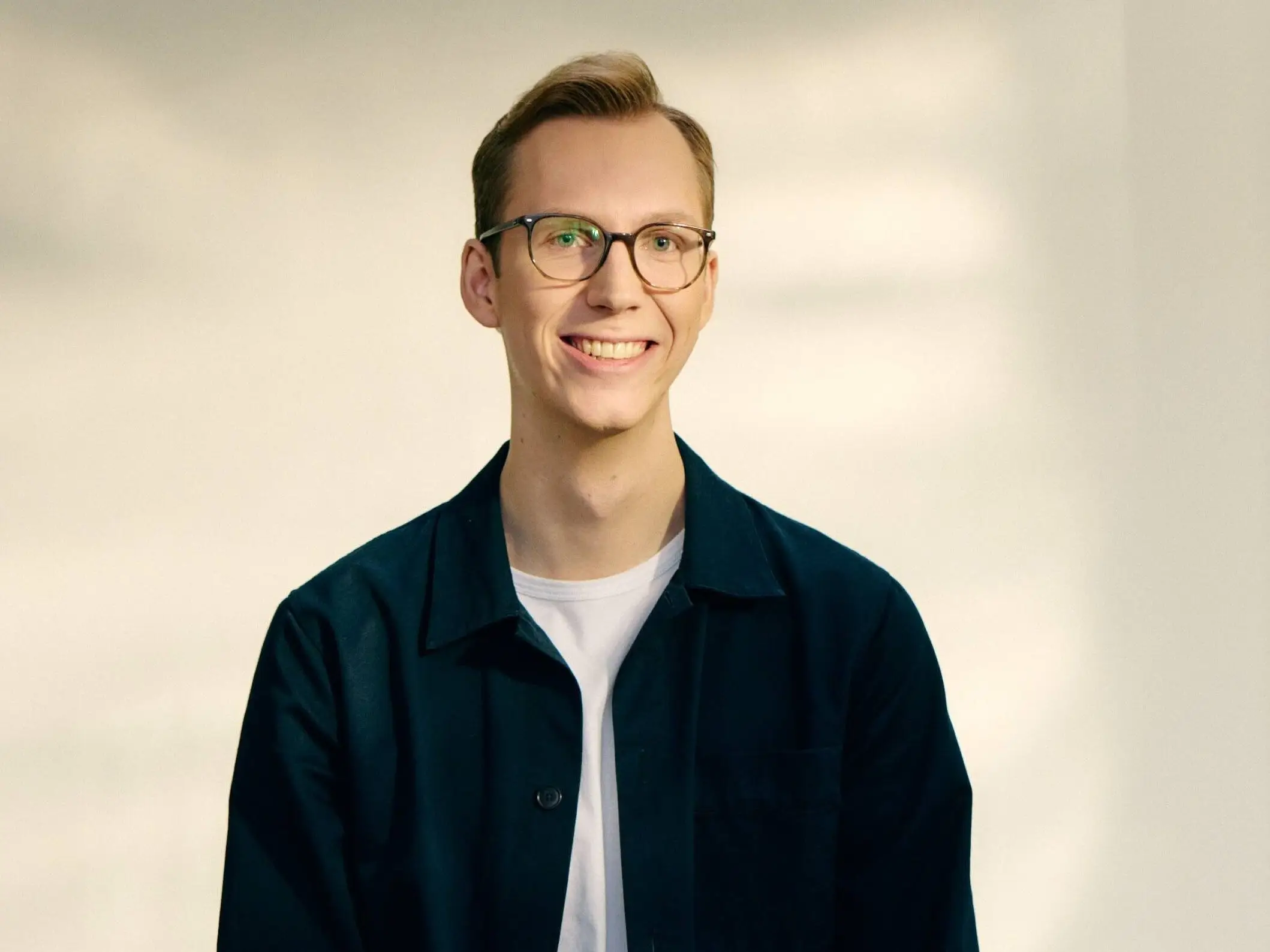 A smiling man wearing glasses sits on a stool in a studio, dressed in a dark jacket over a white T-shirt.
