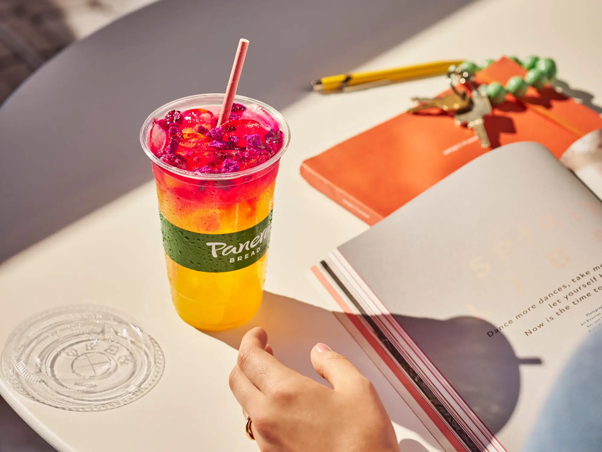 A Panera Energy Refresher sits on a table while a woman reads a book.