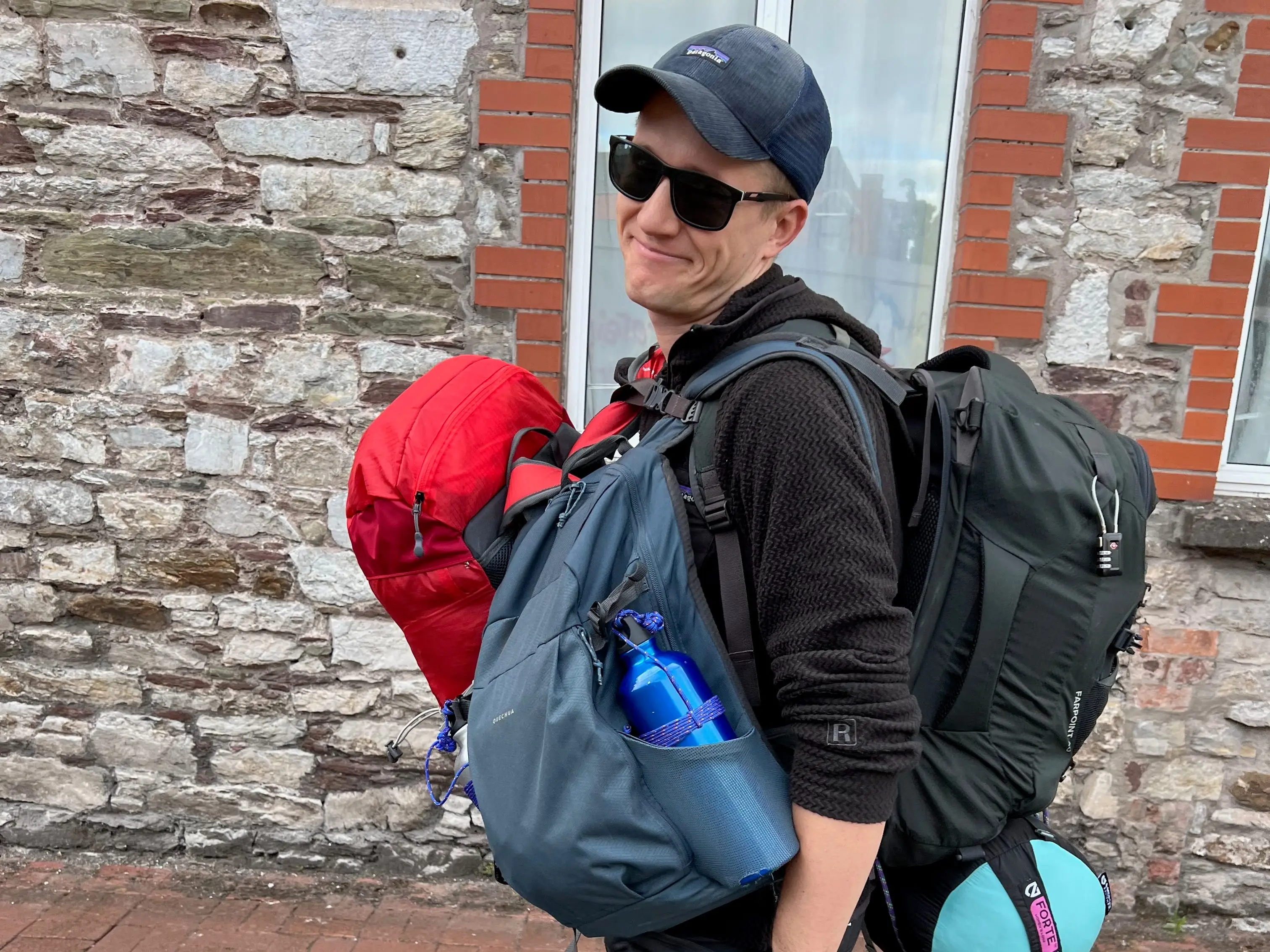 A man wearing a black hat and sunglasses, carrying three large backpacks.
