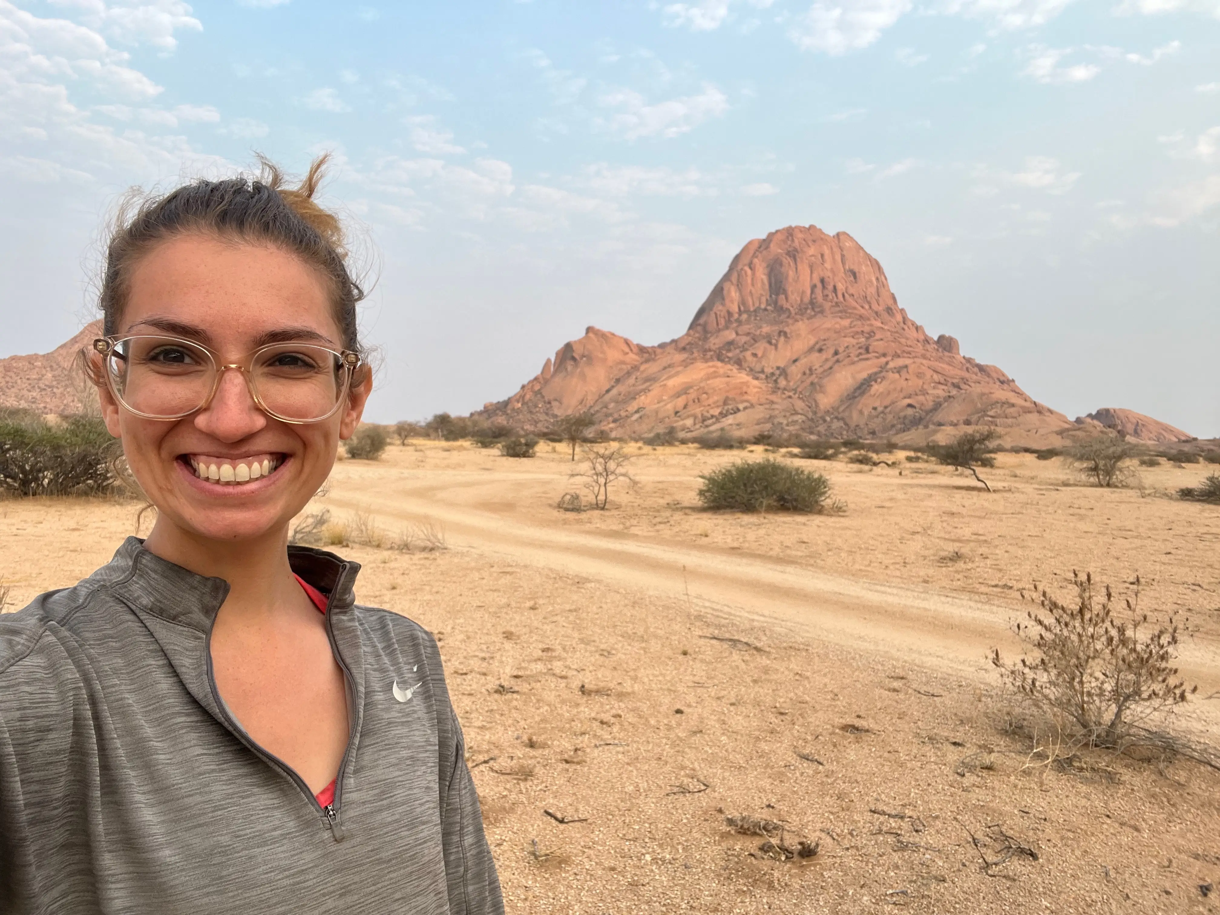 A woman wearing glasses posing with a sandy road and moutain in the background.
