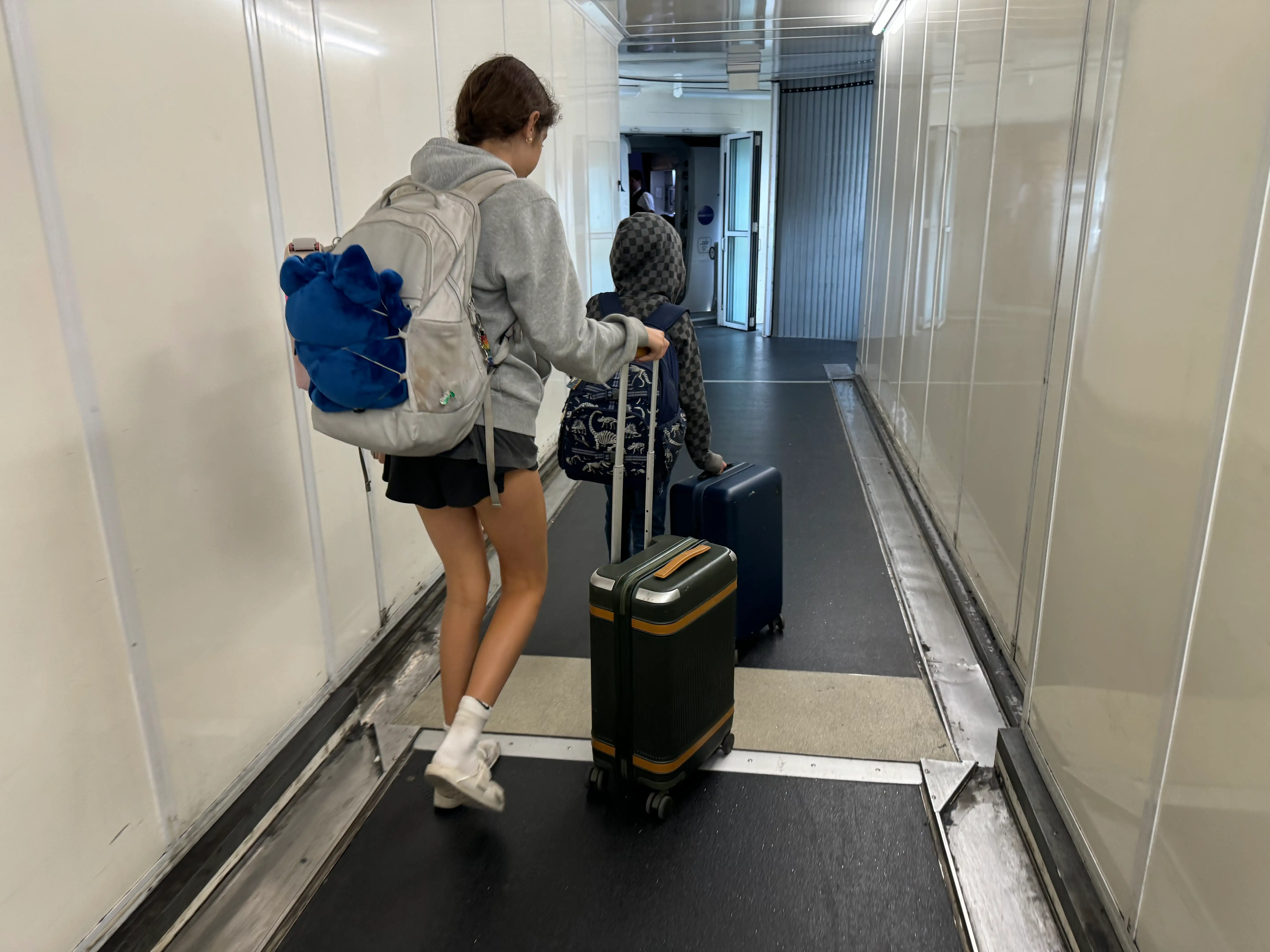 Two kids are shown walking down a jetway with their carry-on bags.