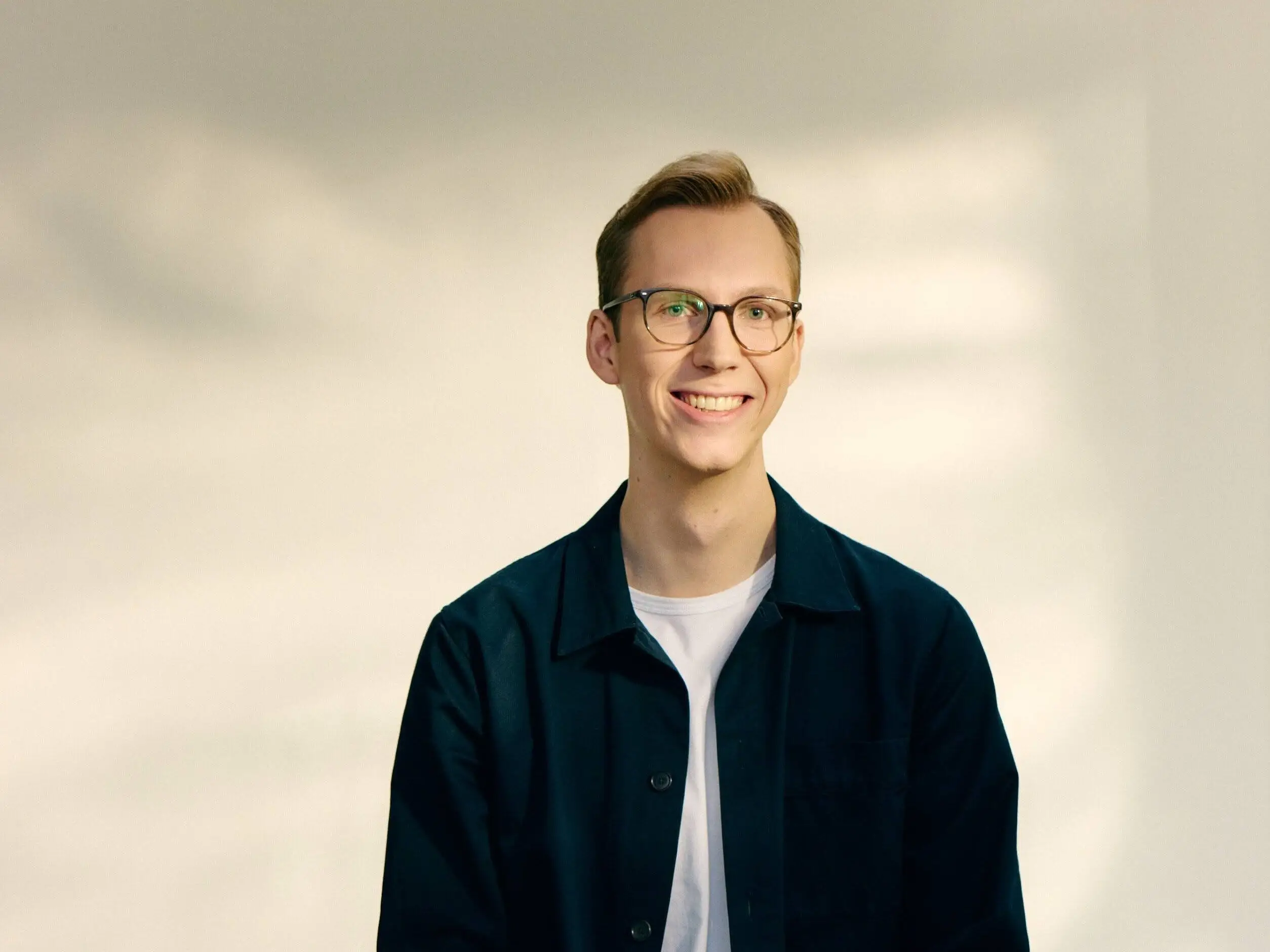 A smiling man wearing glasses sits on a stool in a studio, dressed in a dark jacket over a white T-shirt.