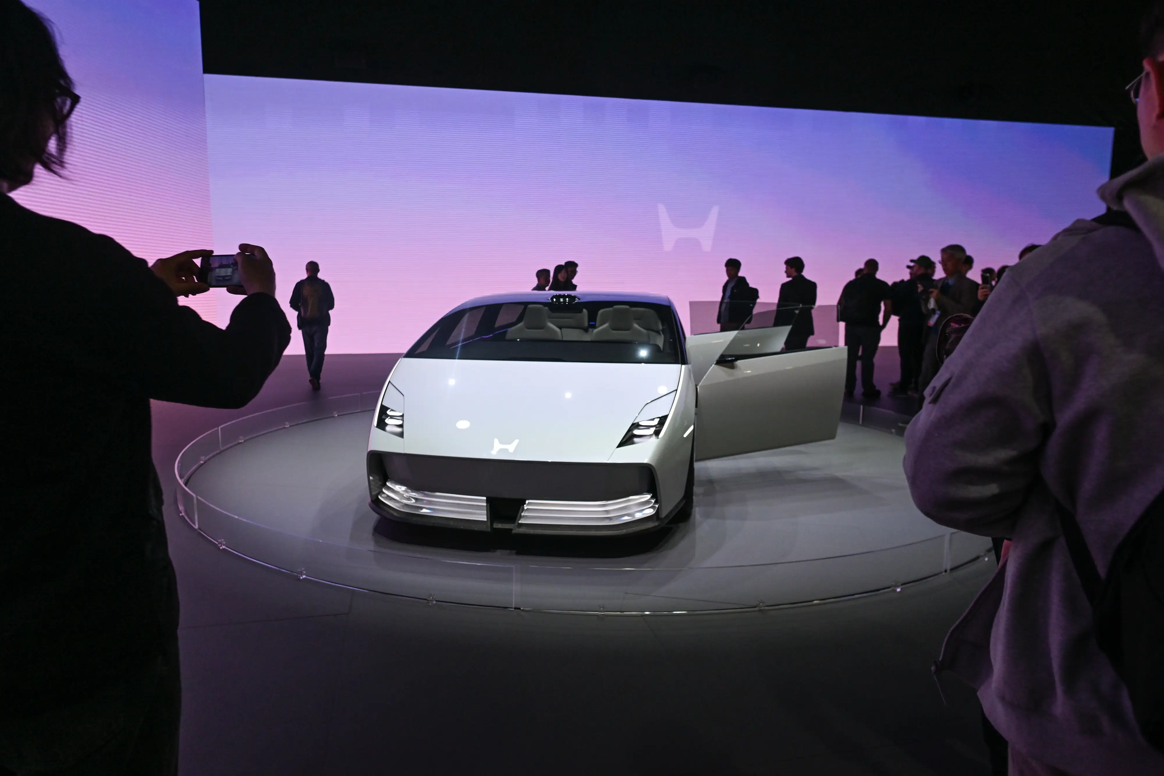 A Honda 0 Saloon is parked on a showroom floor in front of a pink and purple digital screen. Several onlookers are surrounding the car and taking pictures.