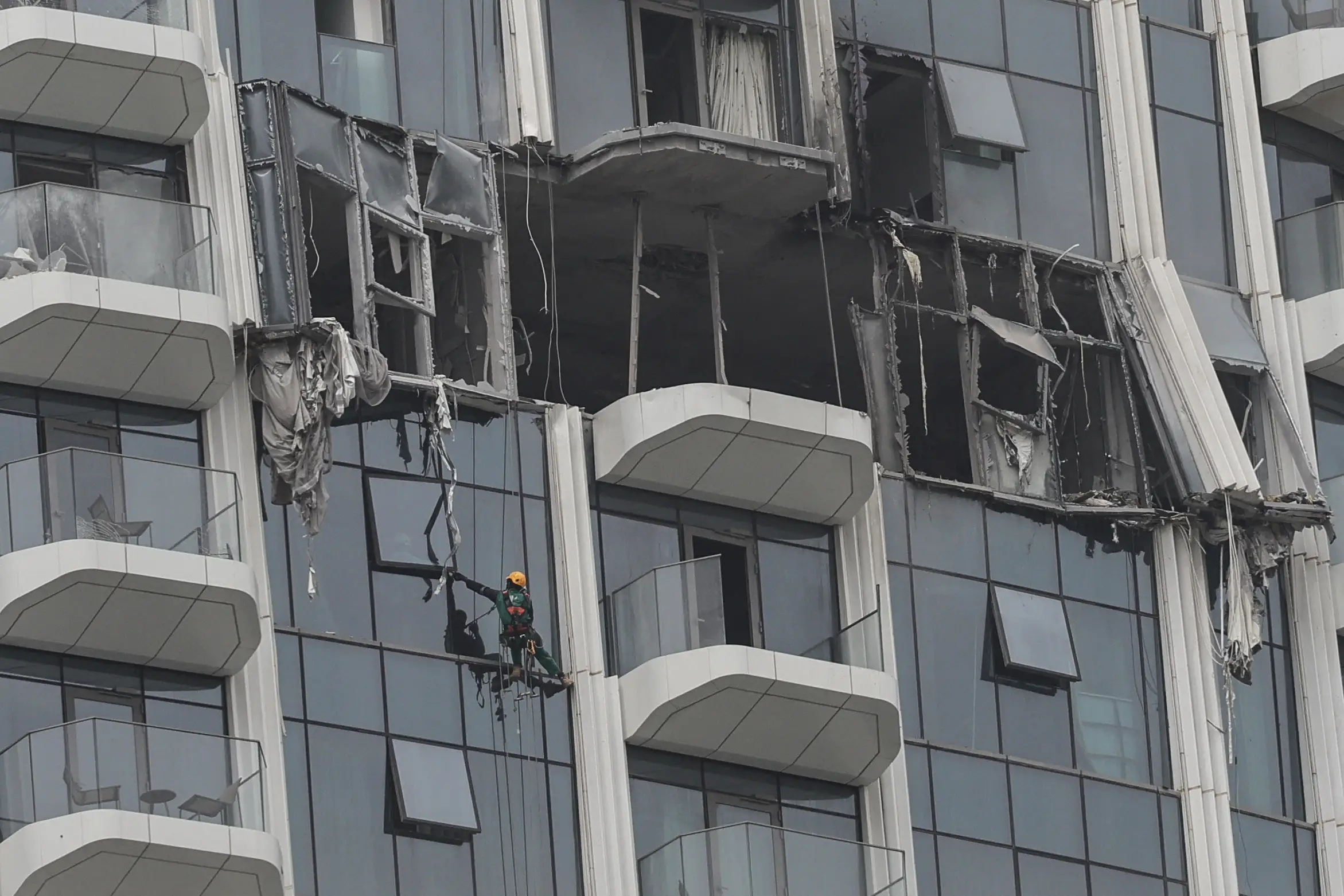 A steeplejack assesses visible damage, including blown-out windows, on the facade of a building.